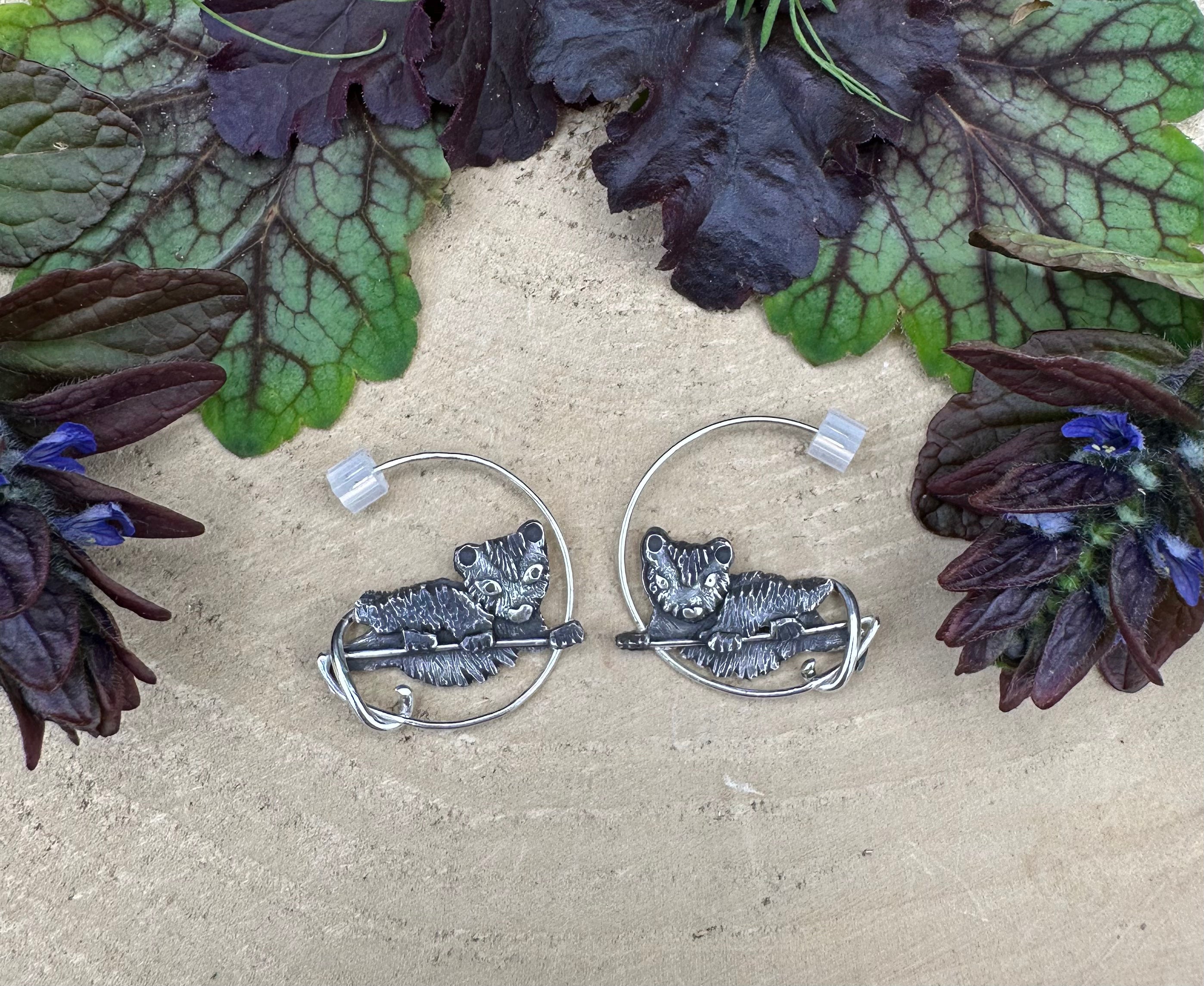 The pair of sterling silver opossum earrings with the silicone backs. The earrings are surrounded by dark purple and green leaves.