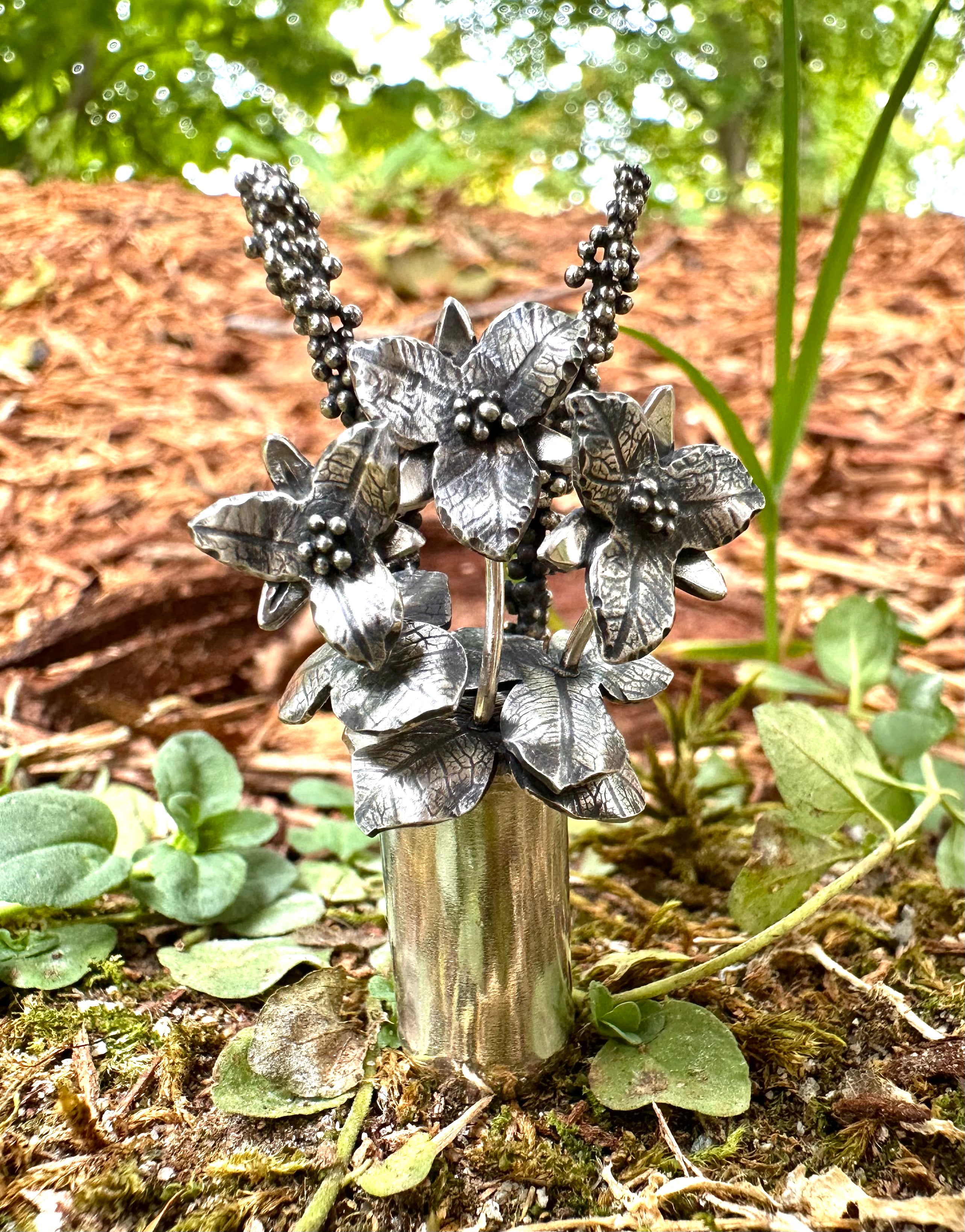 A handmade woman's necklace featuring three trillium flower stems tucked into a vase was made to be worn as a pendant. The pendant is shown on the forest floor.