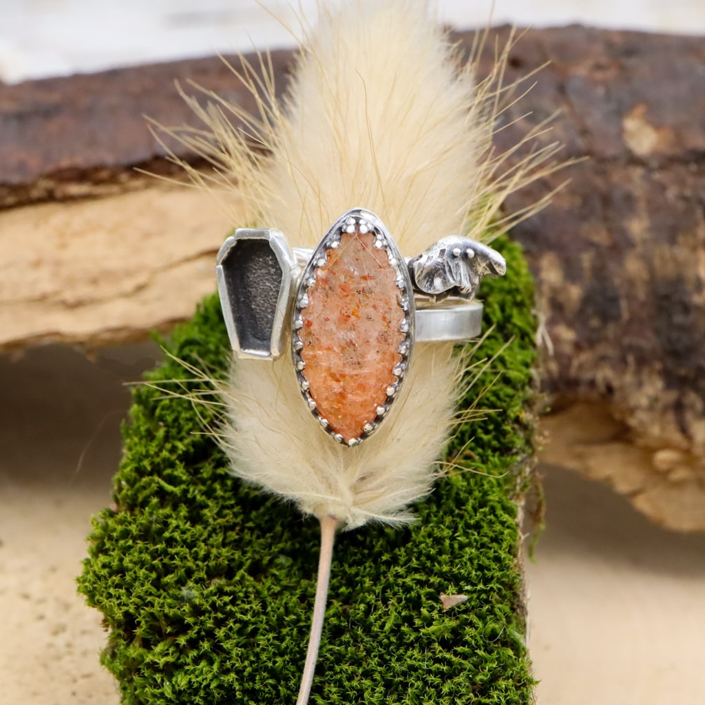 A stack of three rings that are all handmade. There is one band that features a pointed shape rose cut sunstone that is orange, one back has a tiny coffin, and one back has a handmade silver ghost. They are shown on a piece of dark green moss.