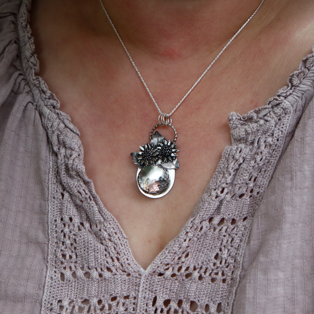 A woman is wearing the tiny mouse and sunflower necklace. She is wearing a light purple blouse.