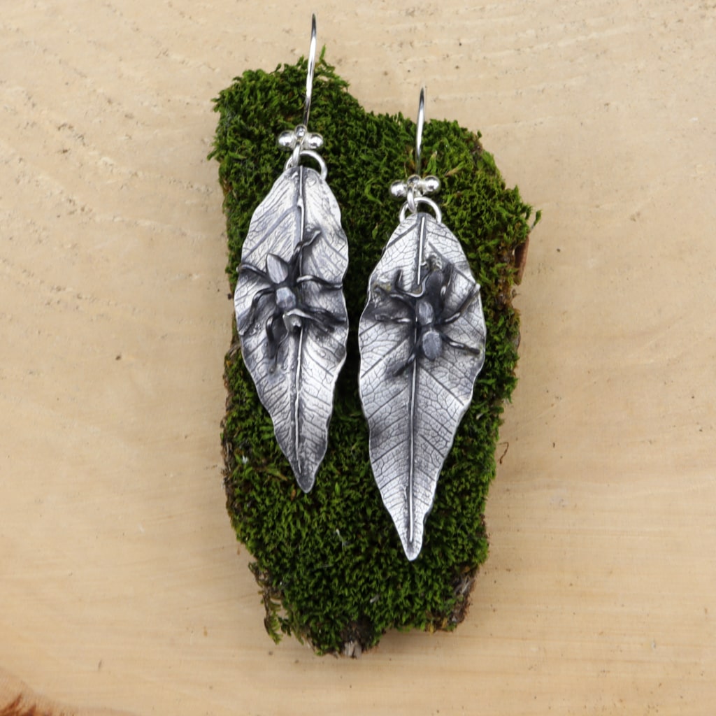 A pair of handmade sterling silver earrings. Each leaf earring has a tiny hand carved ant on them. The earrings are shown on a piece of dark green moss.