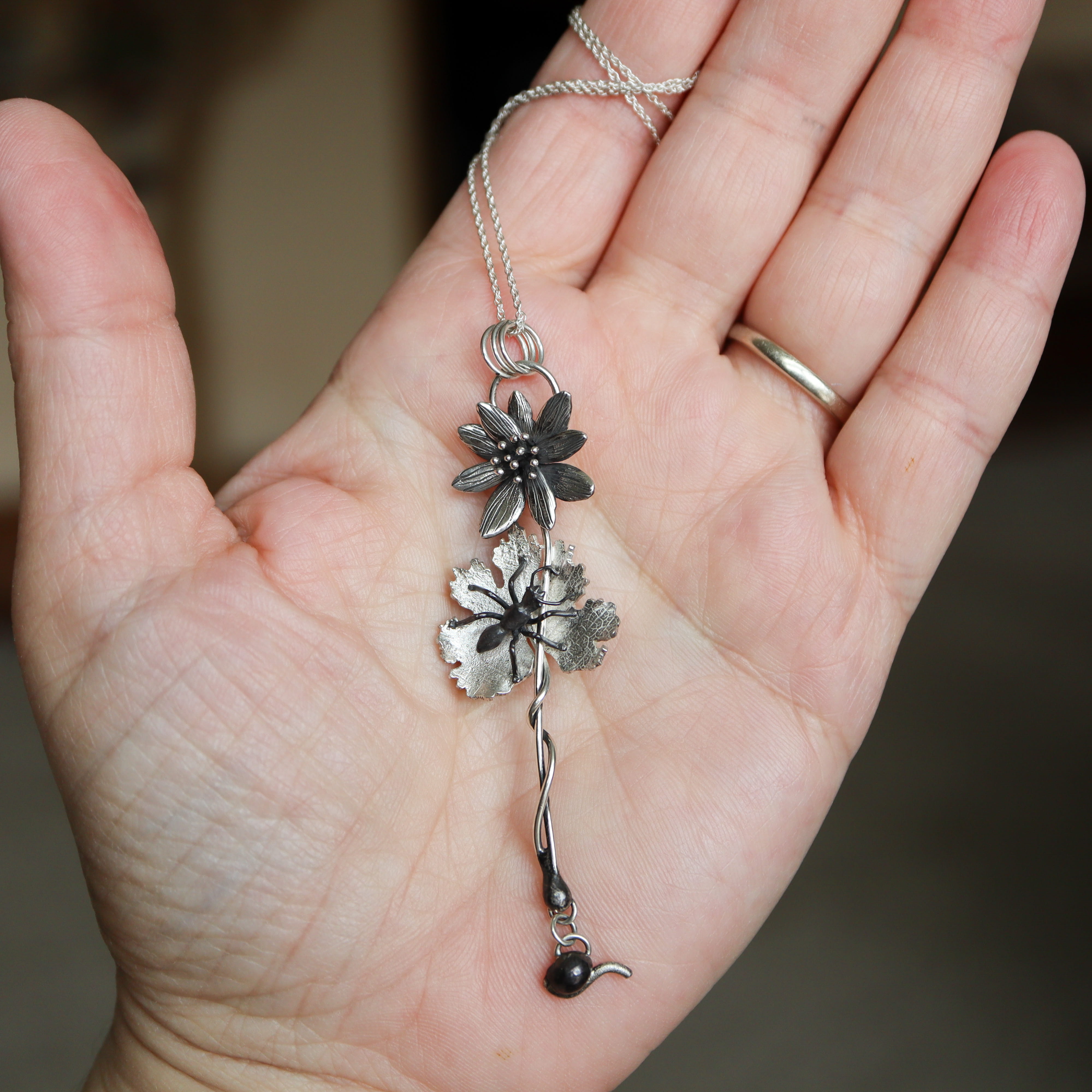 A hand shown holding a handmade sterling silver bloodroot flower necklace that features a tiny ant on the leaf.