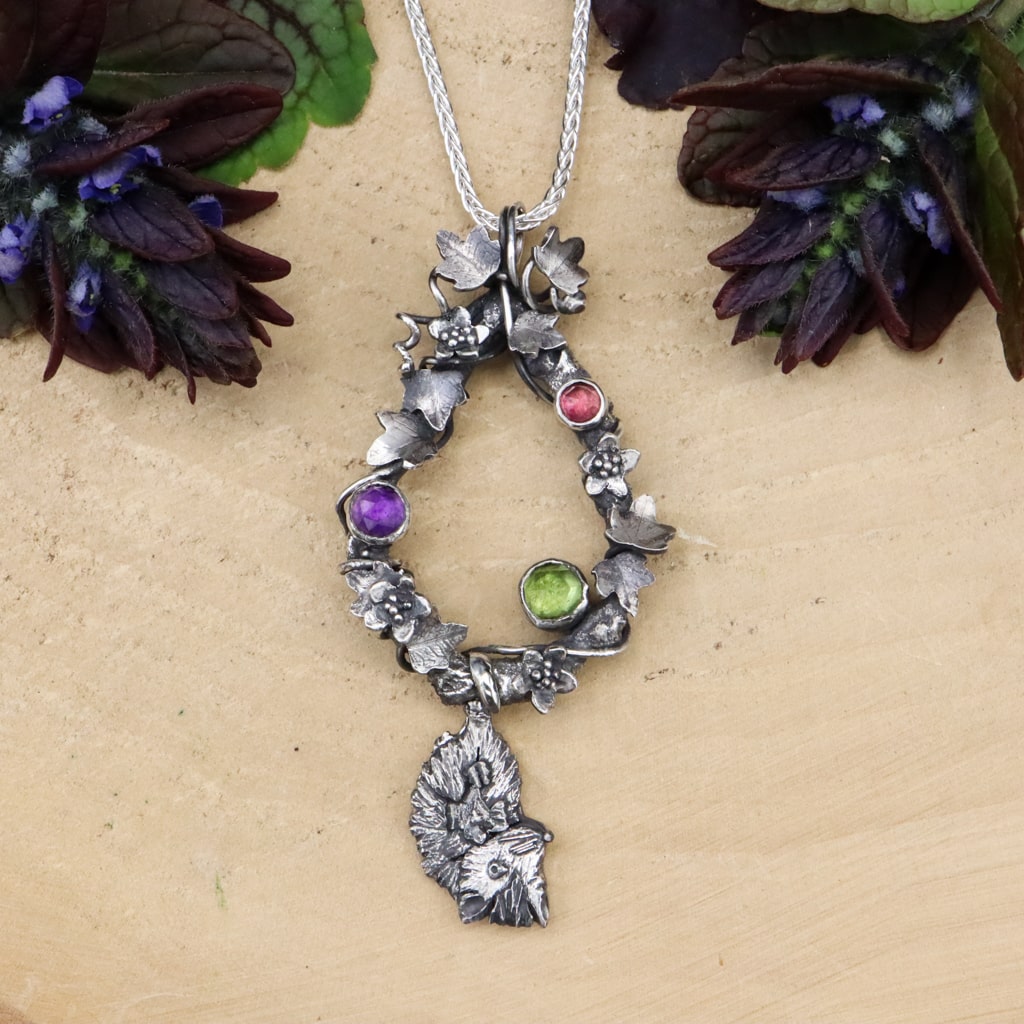 A sterling silver handmade Virginia opossum necklace. The baby opossum is hanging from the bottom of a teardrop shaped tree branch. The branch is covered in tiny ivy leaves and apple blossom flowers. There is a purple amethyst, green peridot, and a pink tourmaline gemstone on the pendant. The pendant is about 3 inches tall and shown on a light tan piece of wood with dark purple flowers on each side of it.