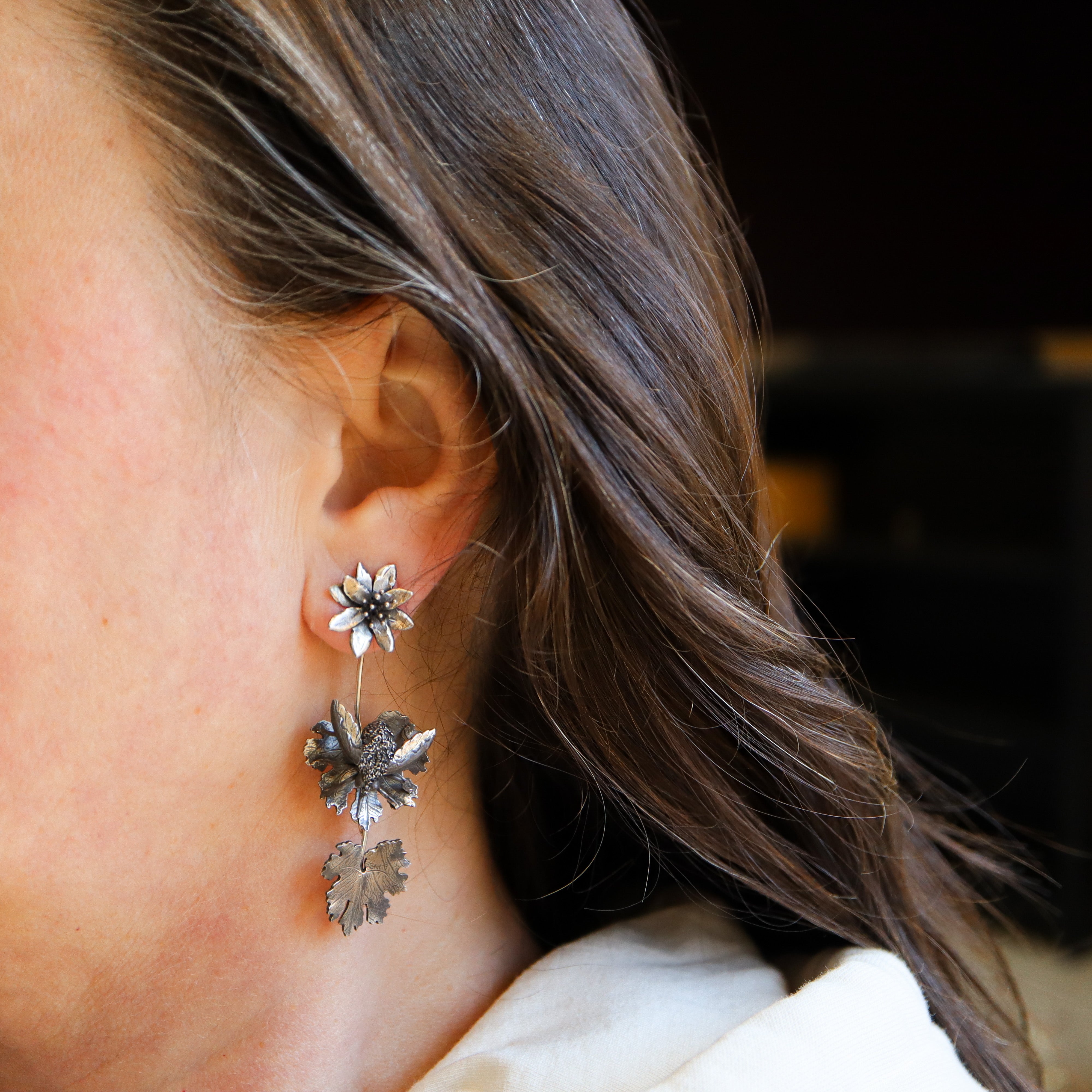 A woman with dark brown hair is shown wearing a pair of handmade sterling silver bloodroot flower and bee fly insect earrings.