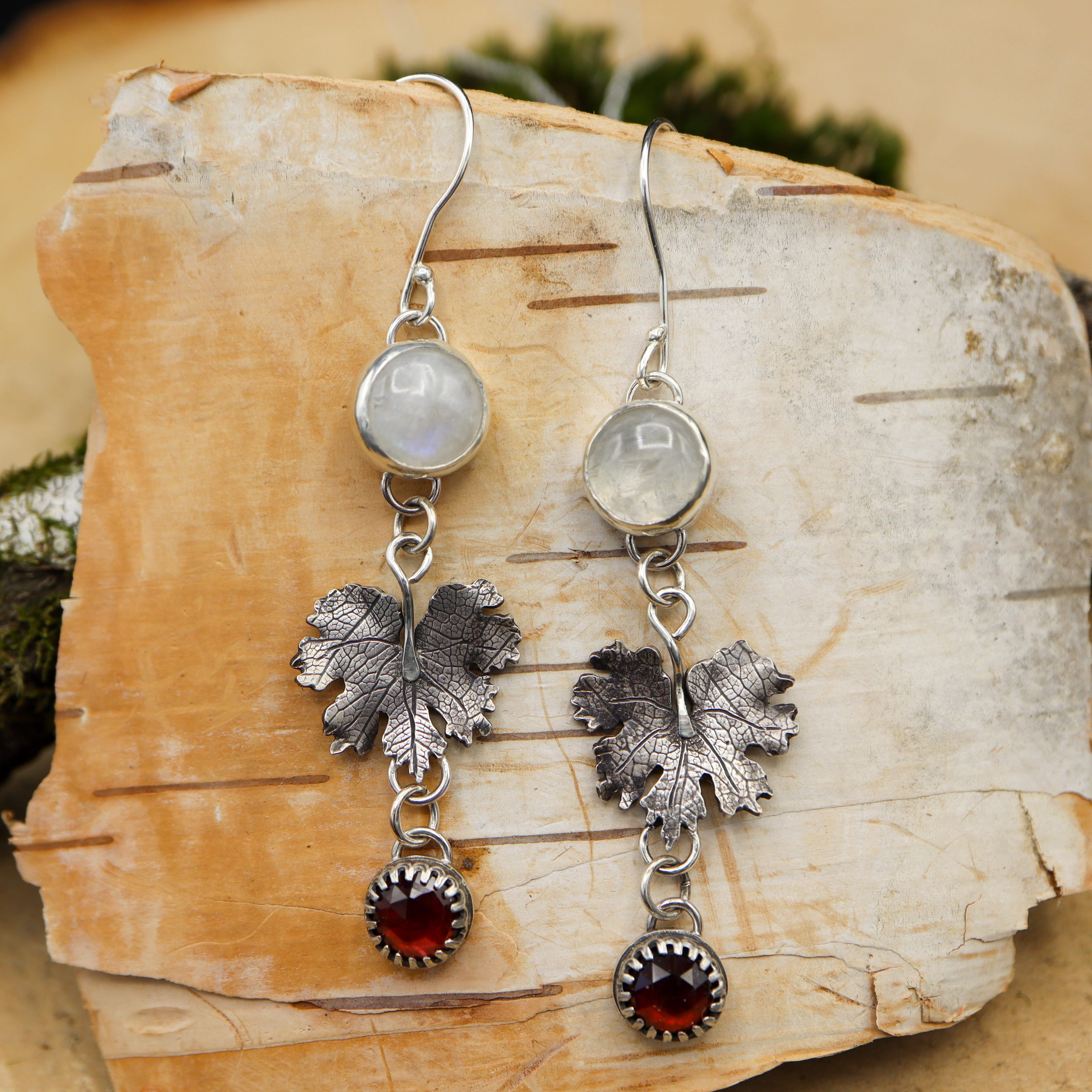 A pair of sterling silver bloodroot earrings with red garnets and moonstone are shown on a piece of birch bark.
