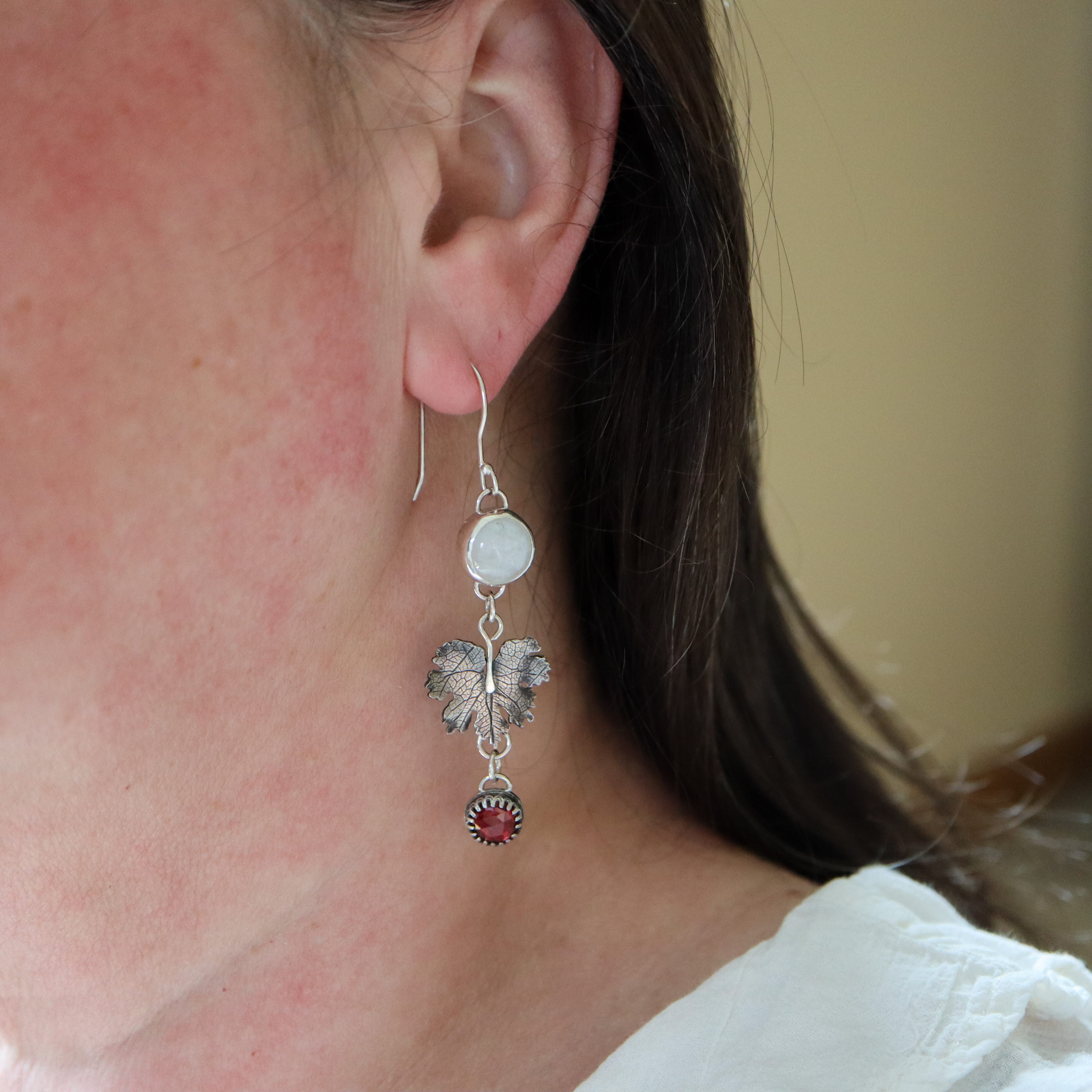 A woman is shown wearing the moonstone and garnet bloodroot leaf flower dangle earrings.