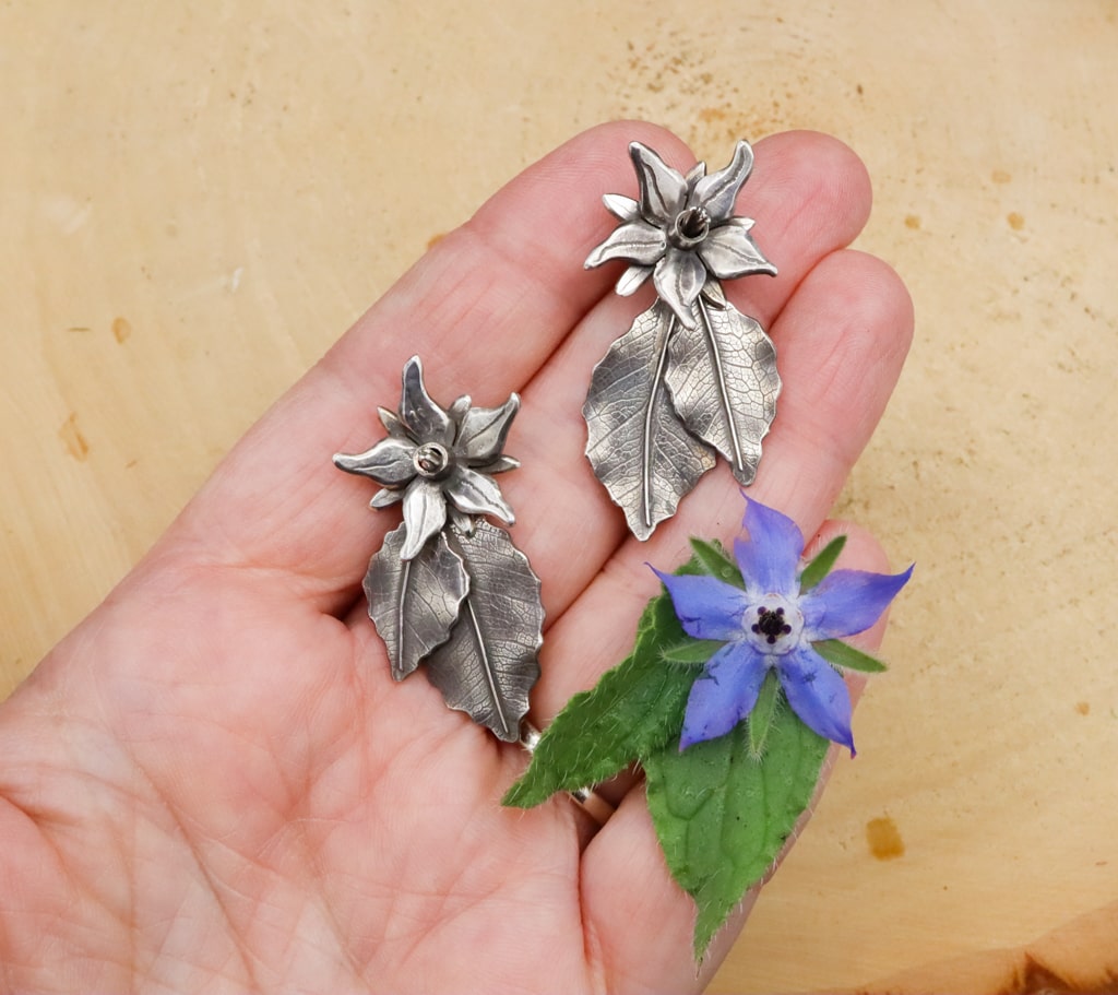 A hand is shown holding both earrings and there is a real borage flower below them to show the size and detail of the earrings.
