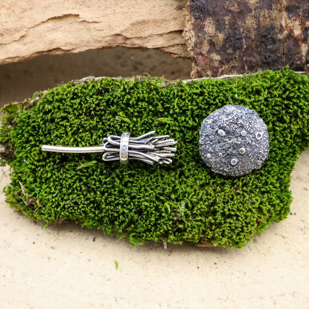 A close up photo of the witches broom and full moon earring studs made from sterling silver. The earring pair are shown on a piece of dark green moss. 