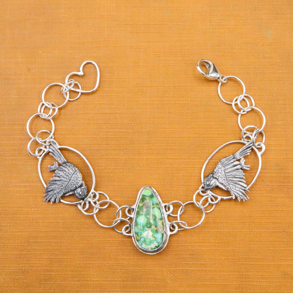 A bracelet featuring two chickadees on either side of a pear shaped Sonoran Mountain turquoise stone. The bracelet is shown on a bright orange vintage book.