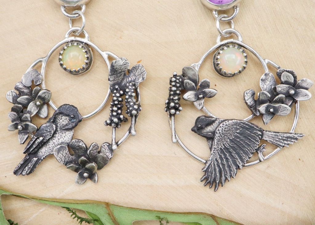 A closer view of the handmade sterling silver chickadee earrings featuring violet flowers, leaves, and hyacinth flowers. They are shown on a piece of light tan wood.