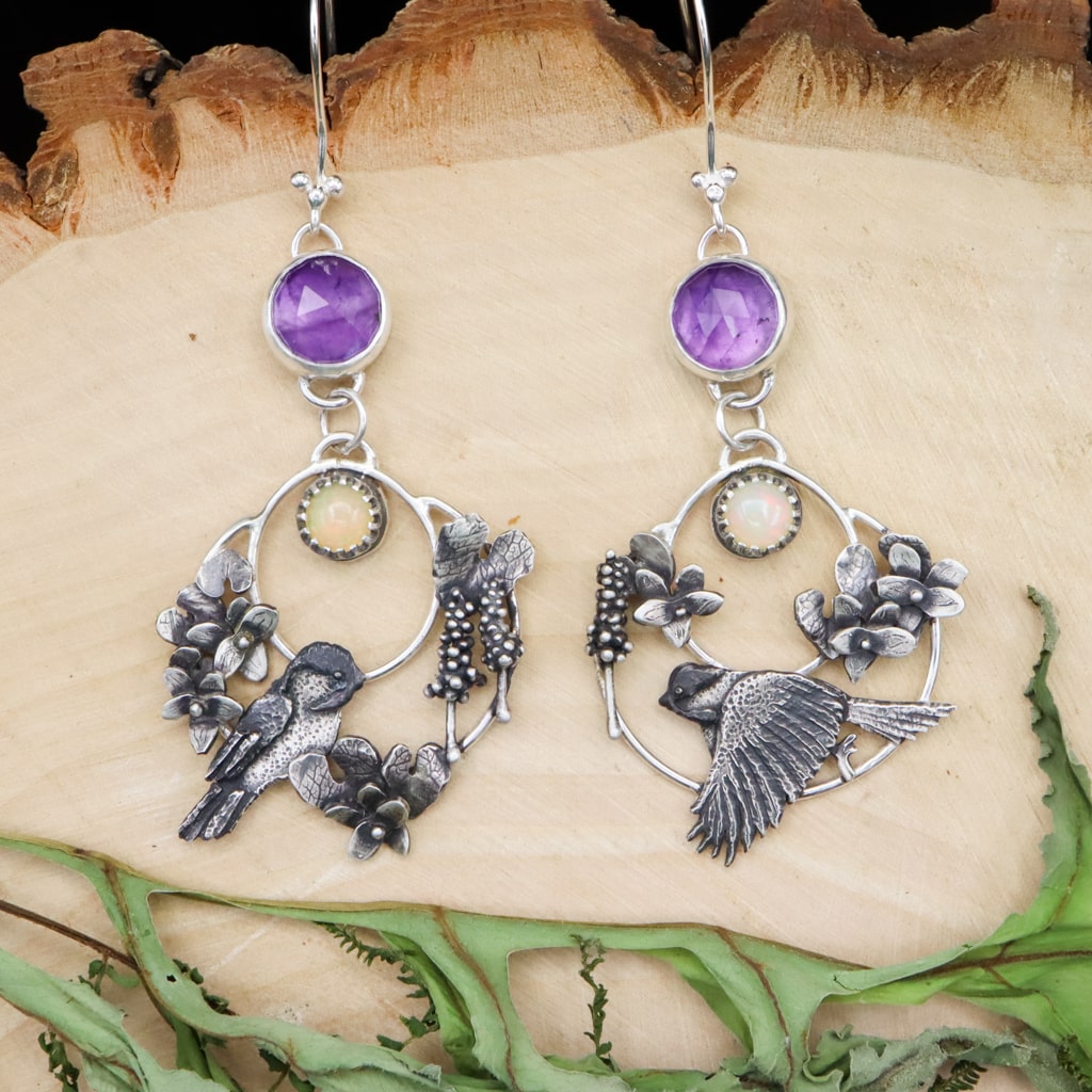 Sterling silver handmade chickadee bird earrings are shaped into hoops. They have an opal stone and amethyst stone at the top. The chickadees are surrounded by tiny violet flowers, leaves, and hyacinth flowers. The earrings are shown on top of a piece of light tan wood.