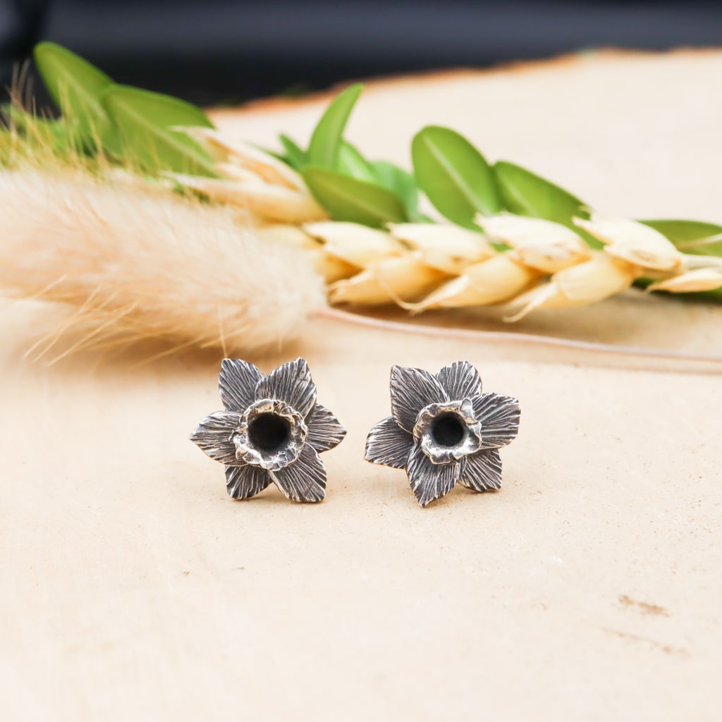 A pair of handmade sterling silver daffodil flower earring studs. They are about 1/4 inch wide and shown on a piece of light tan wood with grasses and greenery in the background.