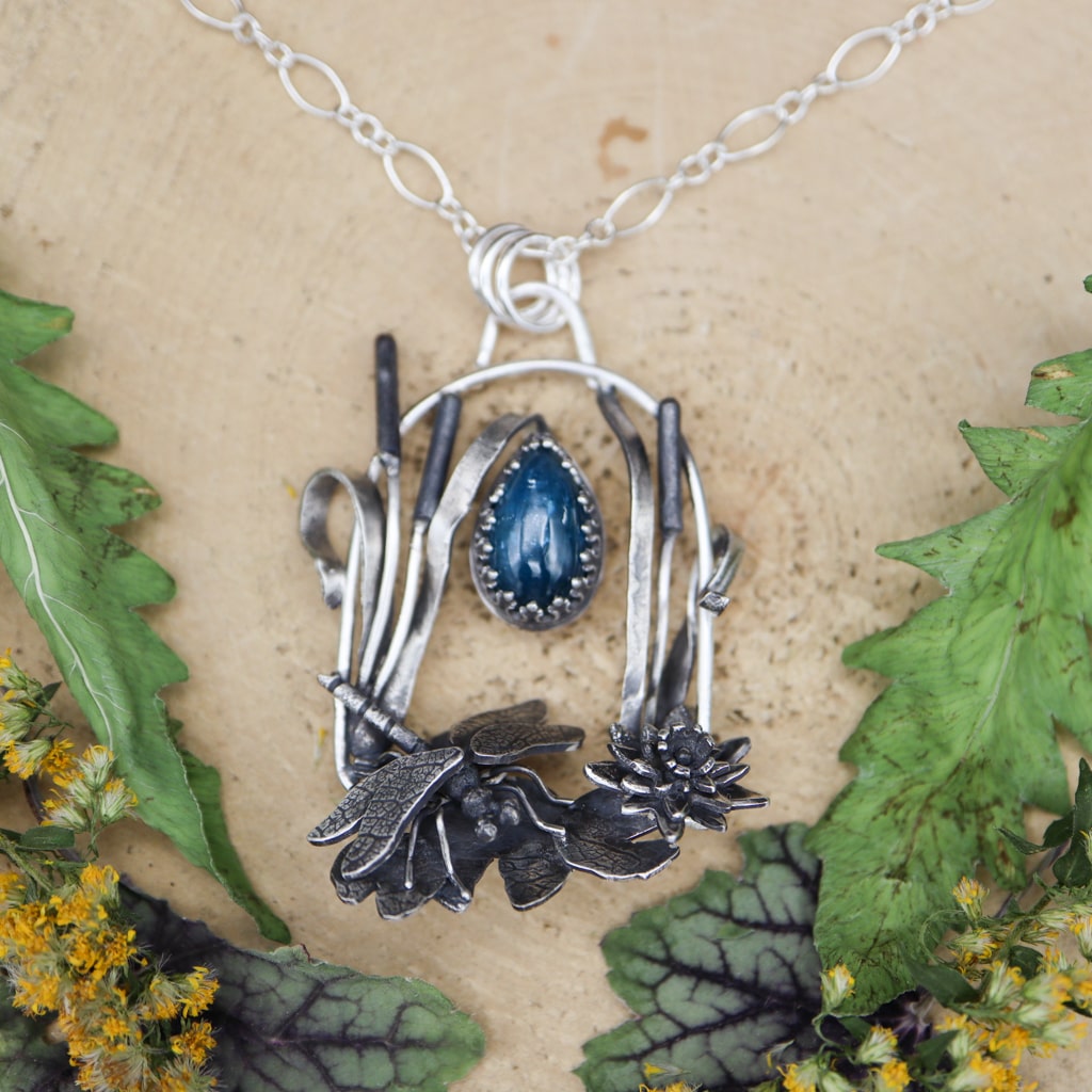 A sterling silver handmade dragonfly necklace is shown among some cattails and lilypads. The necklace also has a deep blue kyanite stone at the top. It is shown on a piece of tan wood and surrounded by dry greens and yellow goldenrod flowers.