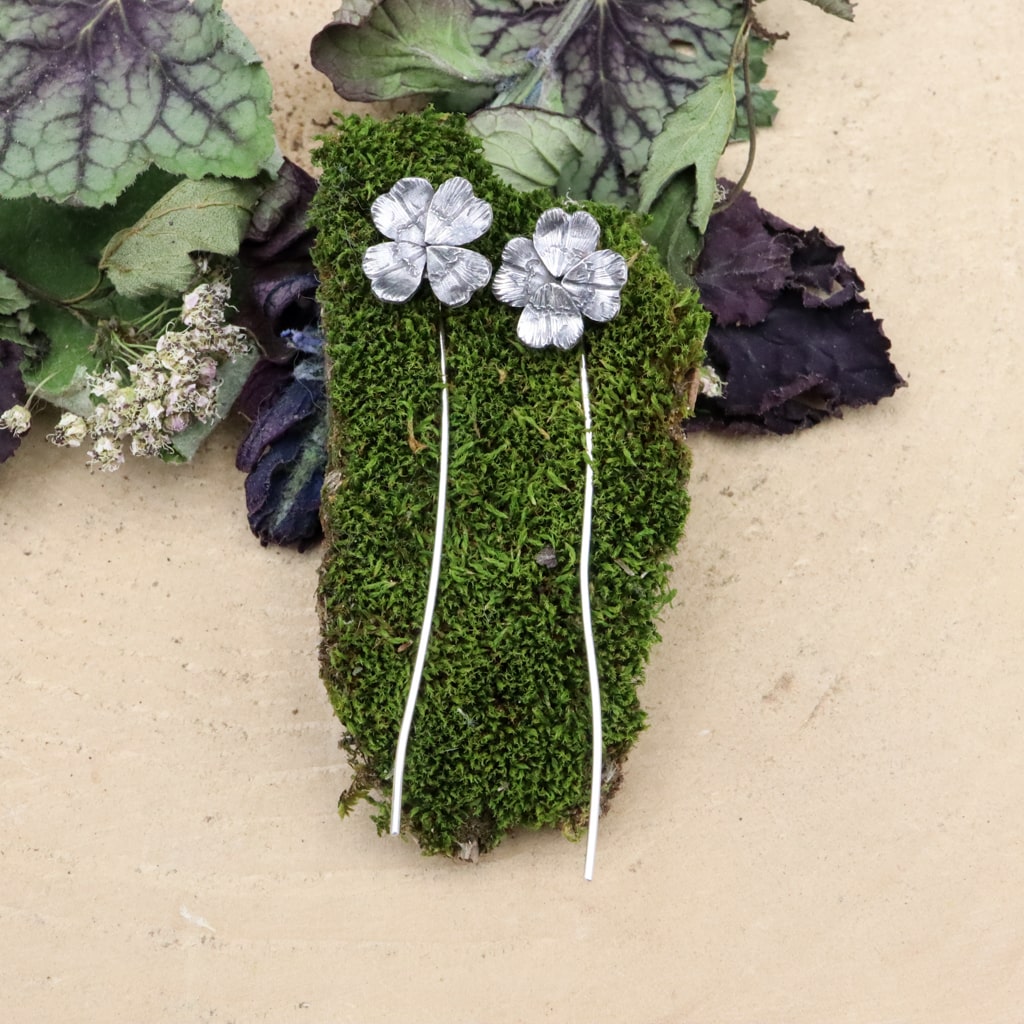 Sterling silver 4 leaf clover earrings are shown on a piece of dark green mos. The clover sits at your earlobe and the stems dangle behind them.