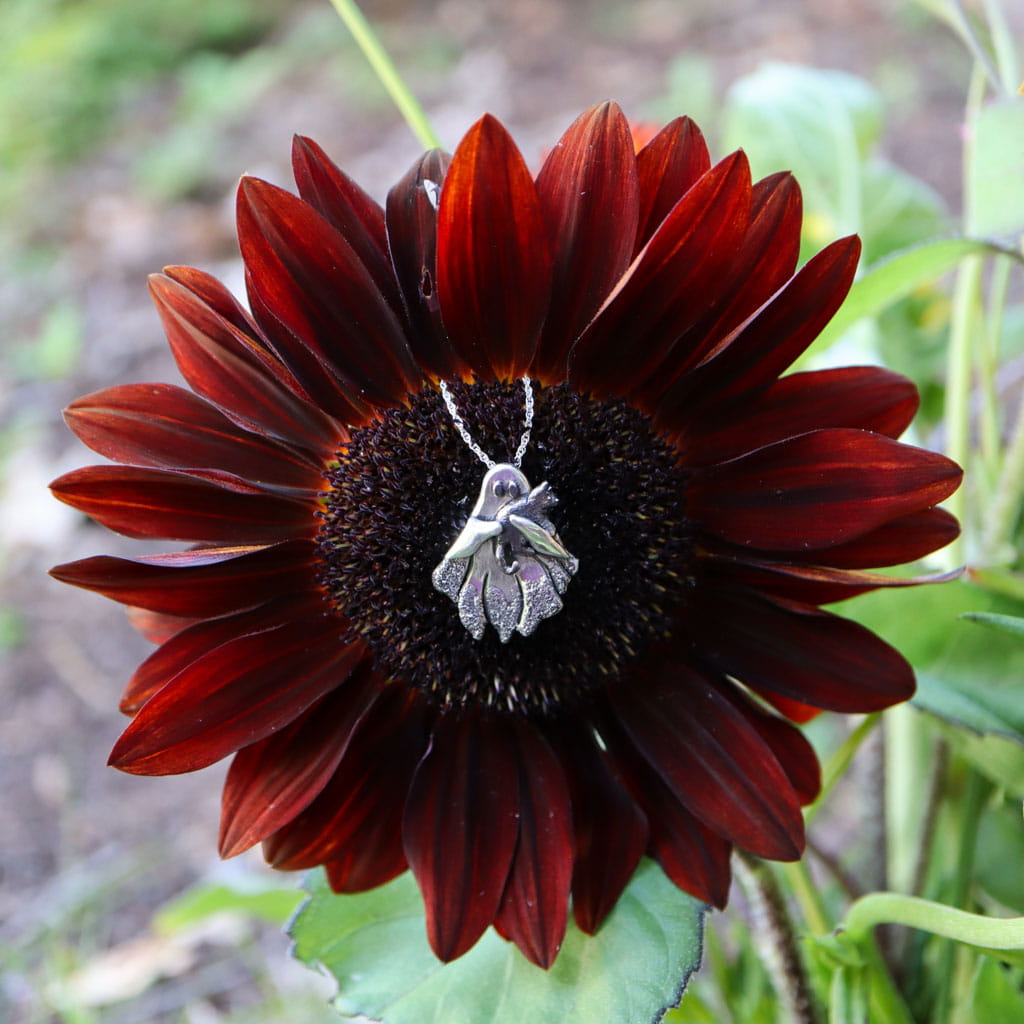 A sterling silver ghost holding a cat pendant necklace hangs from a chain and is placed in the center of a dark red sunflower, with blurred green foliage in the background.