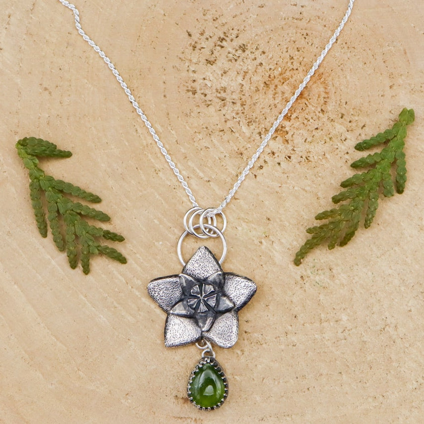 Hoya flower bud necklace is shown on a light tan piece of wood. The flower is about 1 inch tall and below it is a dark green pear shaped vesuvianite stone.