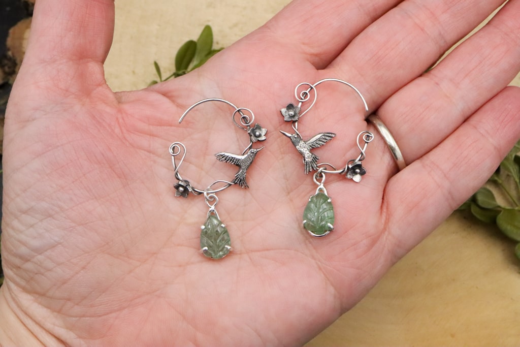 A hand is shown holding a pair of sterling silver handmade hummingbird hoop earrings. They have green dangling leaf gemstones at the bottom and are about 1.75 inches tall.