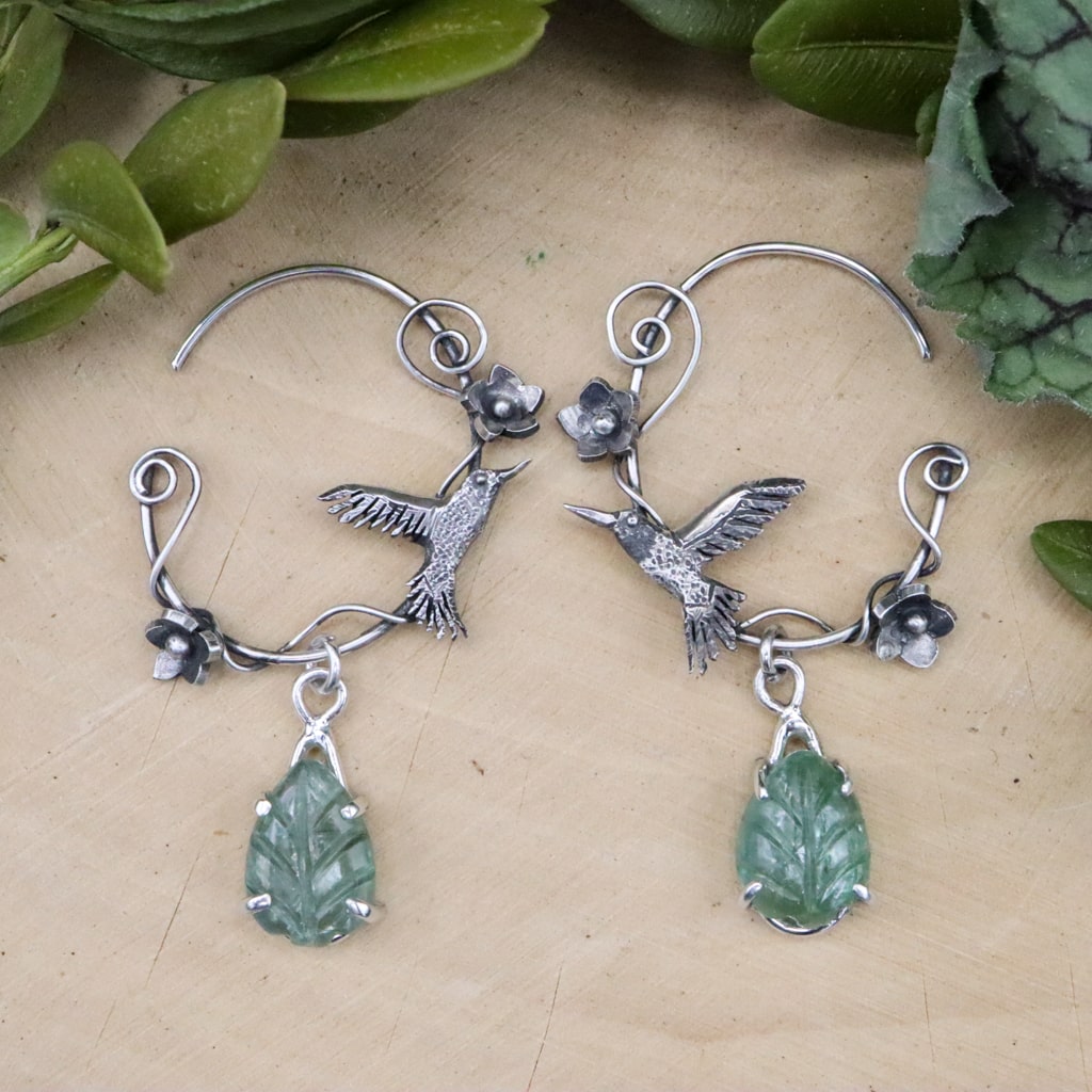 Sterling silver ruby-throated hummingbird hoop earrings are shown on a piece of light tan wood. They are about 1.75 inches long and feature a light green kyanite leaf gemstone below each earring.
