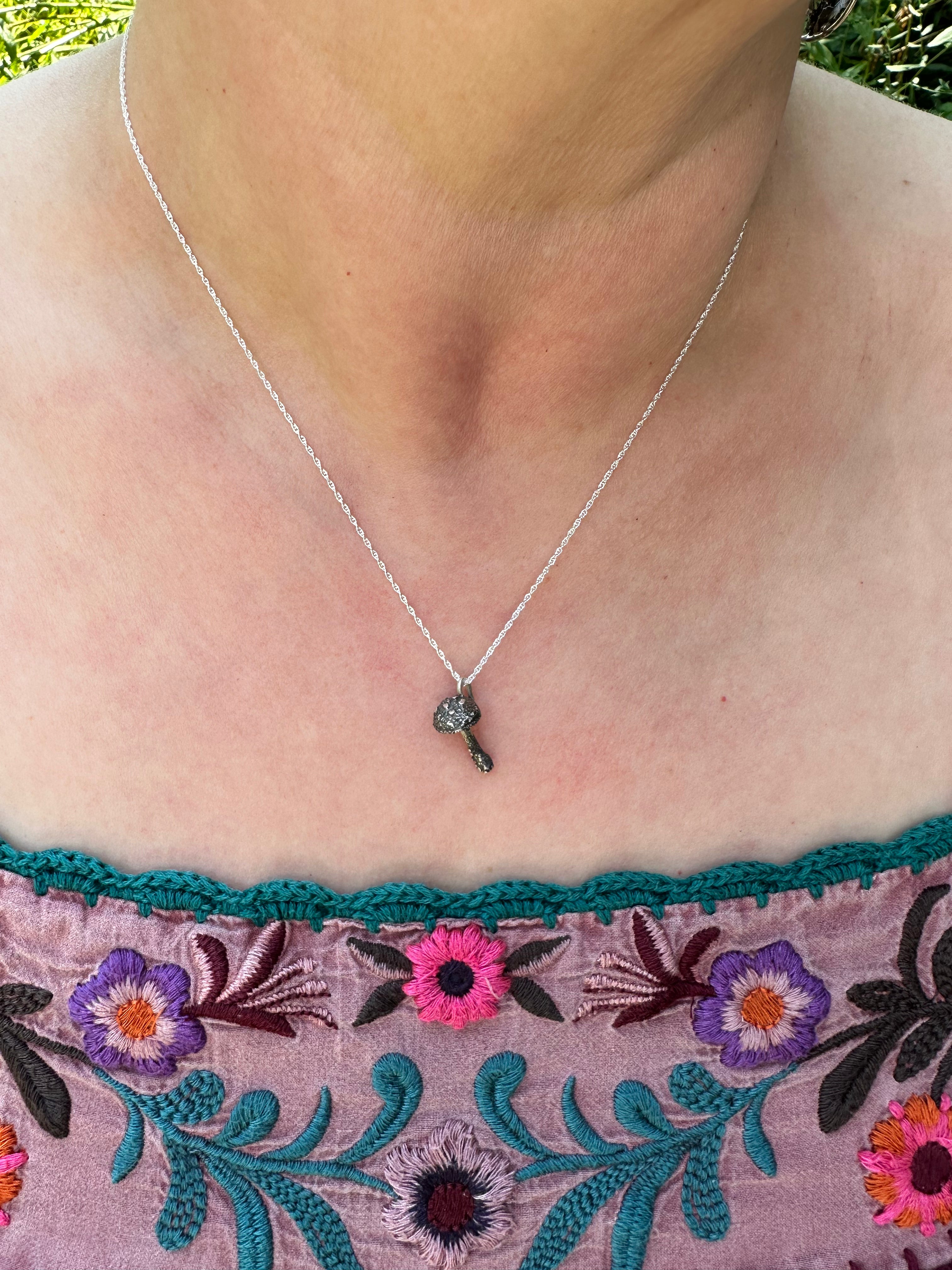 The dainty mushroom necklace is shown being worn by someone with a light pink floral embroidered shirt.