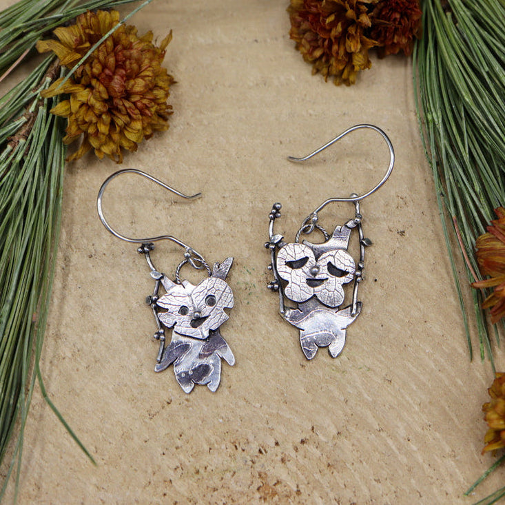 A paid of handmade sterling silver korok earrings each holding a berry twig to shake. they are shown on a piece of light tan wood surrounded by pine needles and flowers.