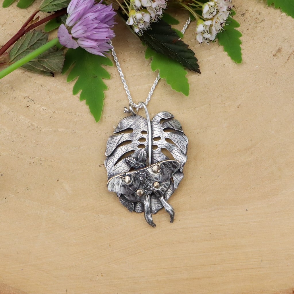 A handmade sterling silver monstera leaf with a silver and gold luna moth sitting upon it. The leaf is attached to a silver necklace and shown on a piece of light tan wood with some white and purple flowers above it.
