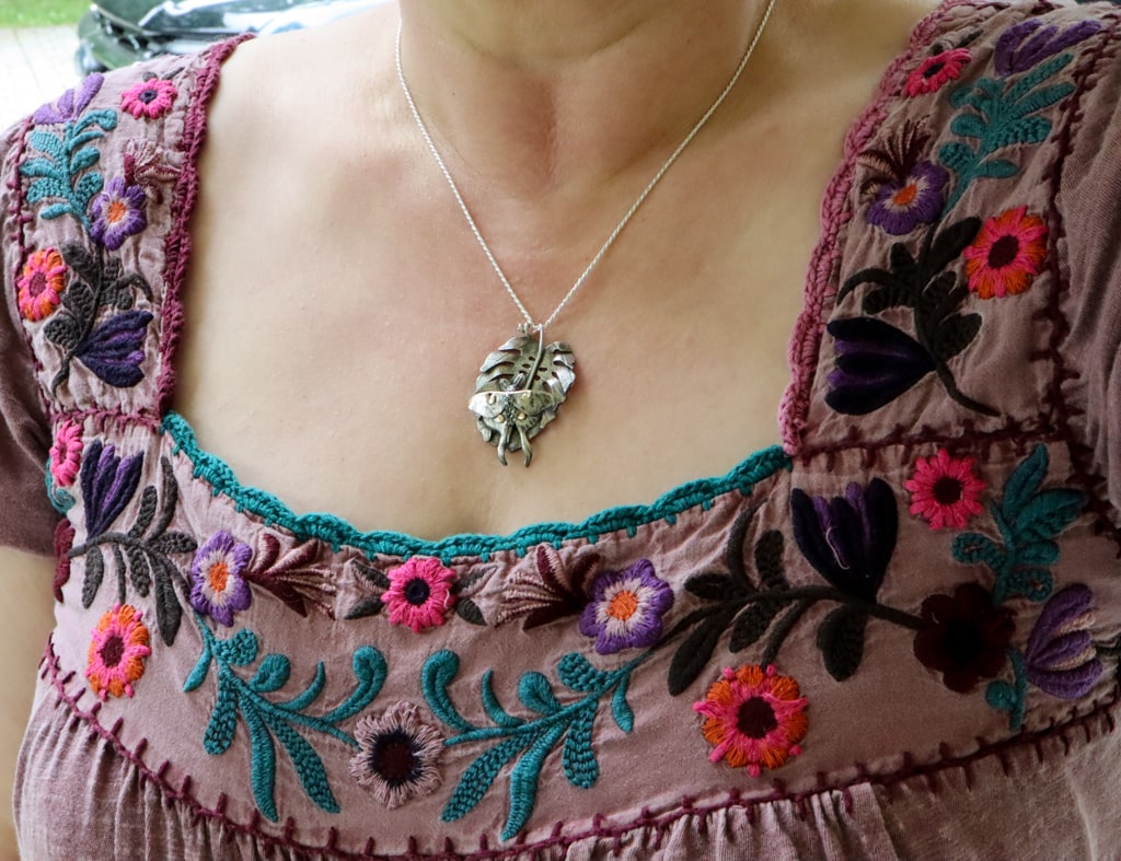 A woman is shown wearing the luna moth and monstera deliciosa leaf necklace being worn. Her shirt is covered in embroidered flowers and a rainbow of colors.