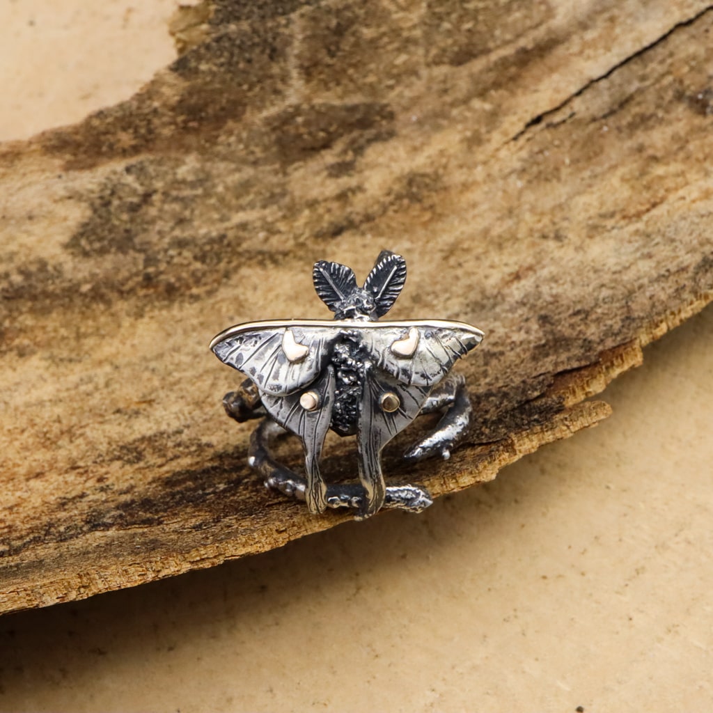 A handmade sterling silver and gold luna moth sits on top of a tree branch ring band. The ring is shown on a piece of light tan wood.