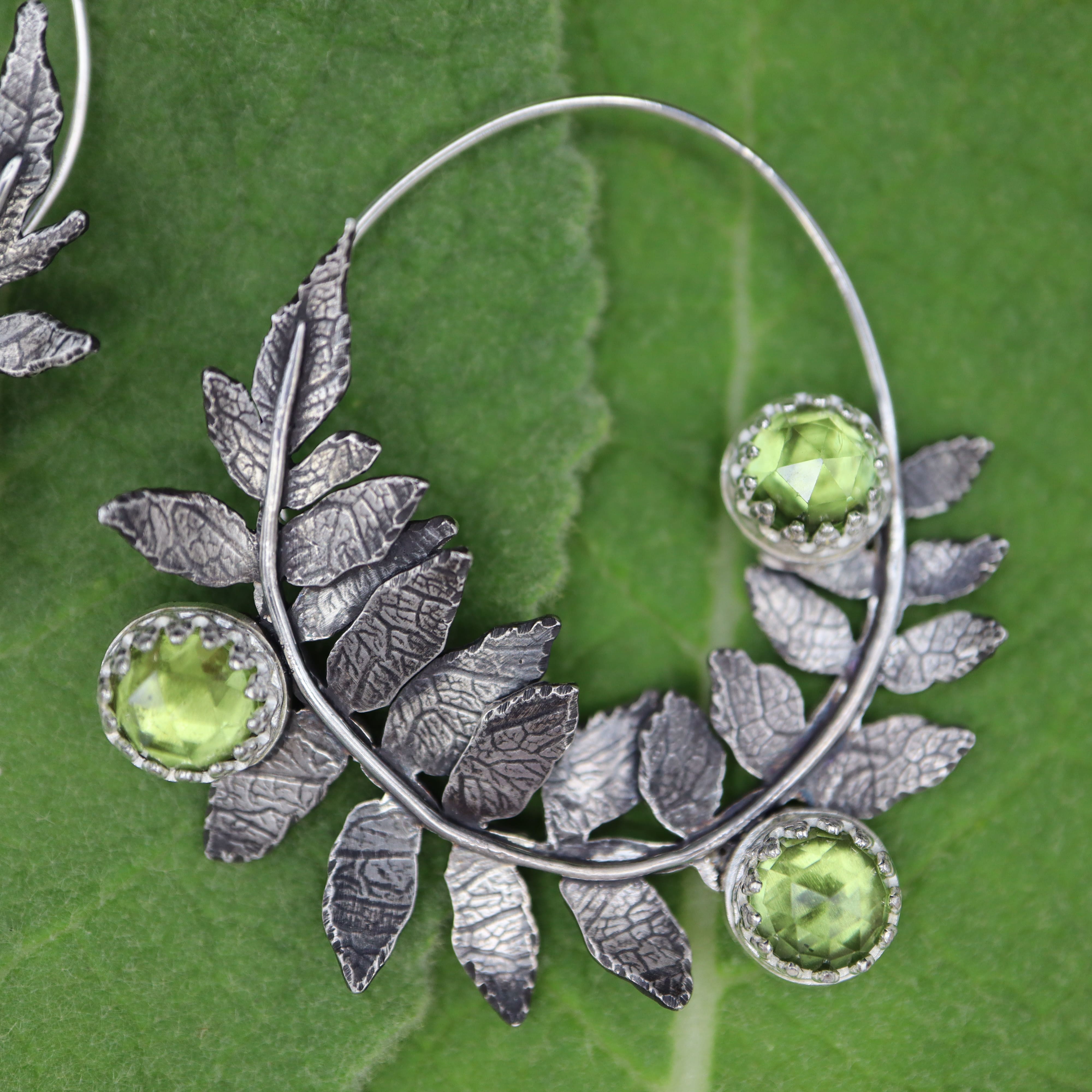 Silver staghorn sumac leaf-shaped earrings with green peridot gemstones on a green leaf background