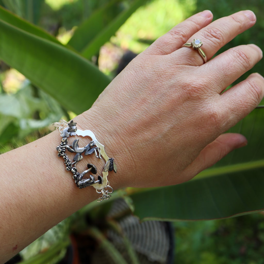 The orchid mantis bracelet is shown being worn on a wrist. There are light green plants in the background.
