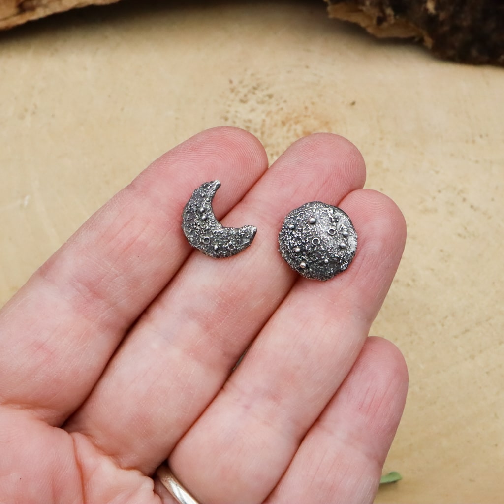 A hand is shown holding the mismatched pair of sterling silver moon phase earrings.