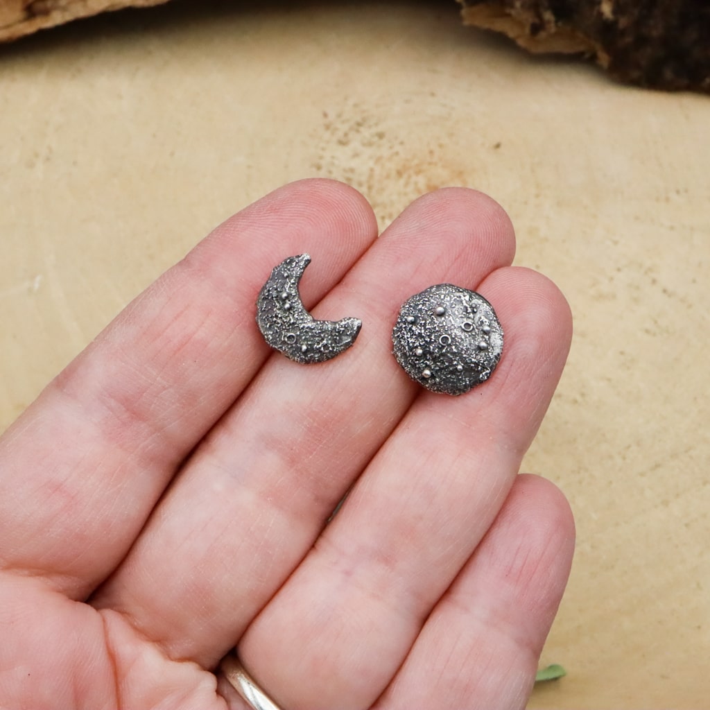 A hand is shown holding the mismatched pair of sterling silver moon phase earrings. 