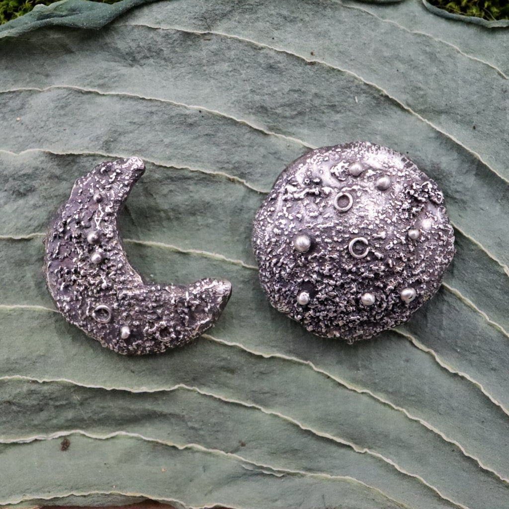 Sterling silver mismatched moon earring studs. One earring is a crescent moon and one earring is a full moon. They are shown on a dark green leaf. 