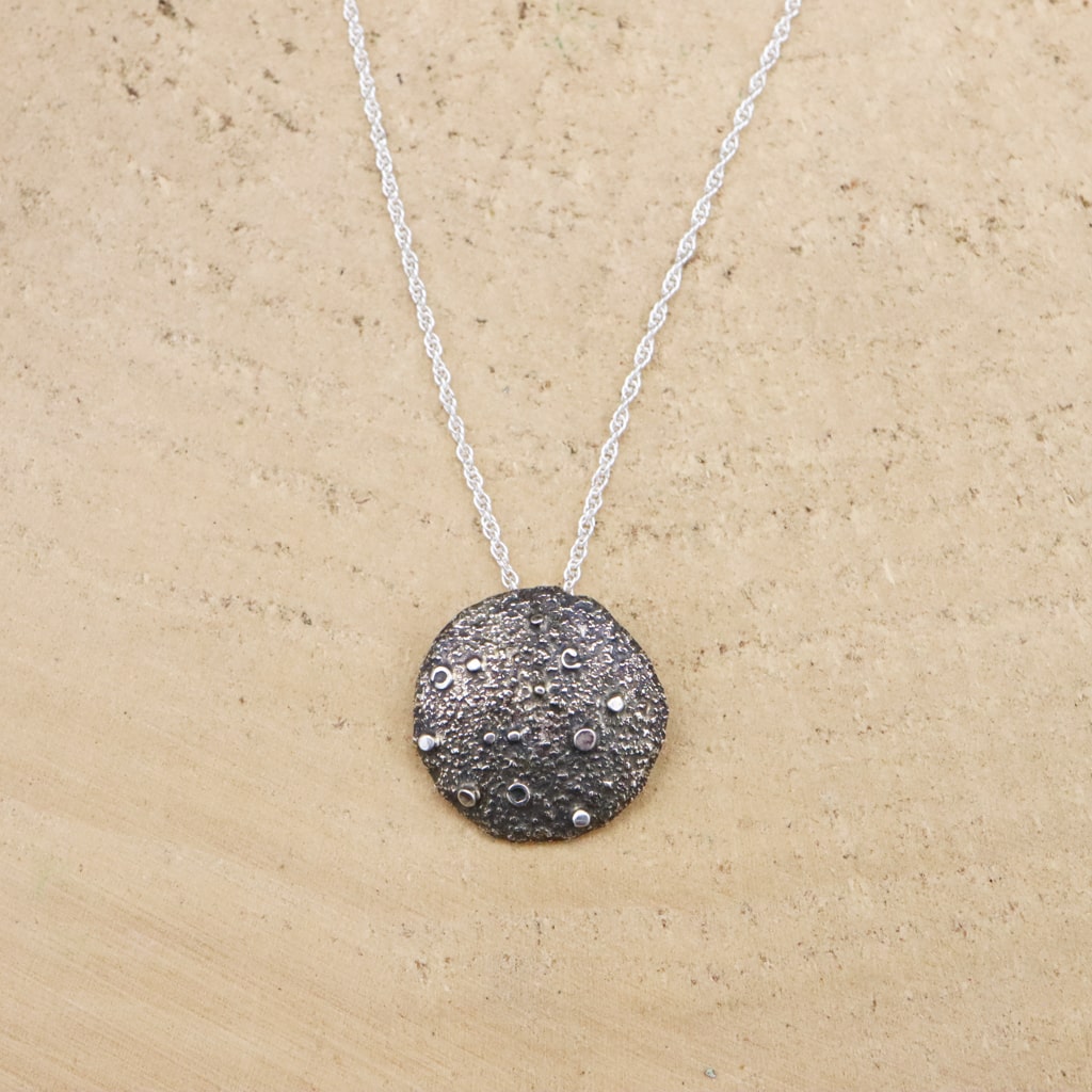 A sterling silver full moon necklace is shown on a light tan piece of wood. The moon is about 1 inch across.