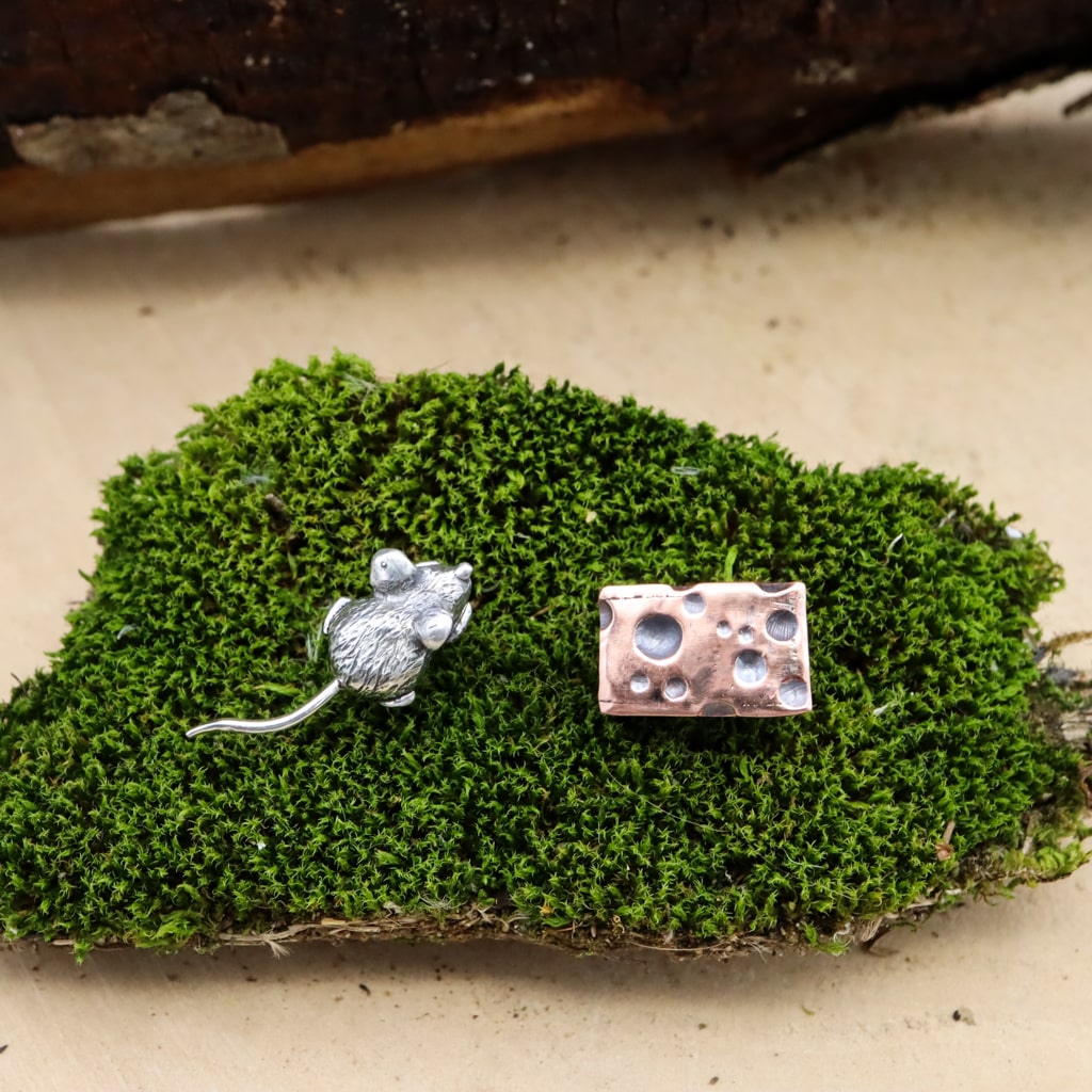 A tiny hand carved sterling silver mouse and a piece of 3 dimensional swiss cheese made from copper. They are a pair of mismatched stud earrings and shown on a piece of green moss.