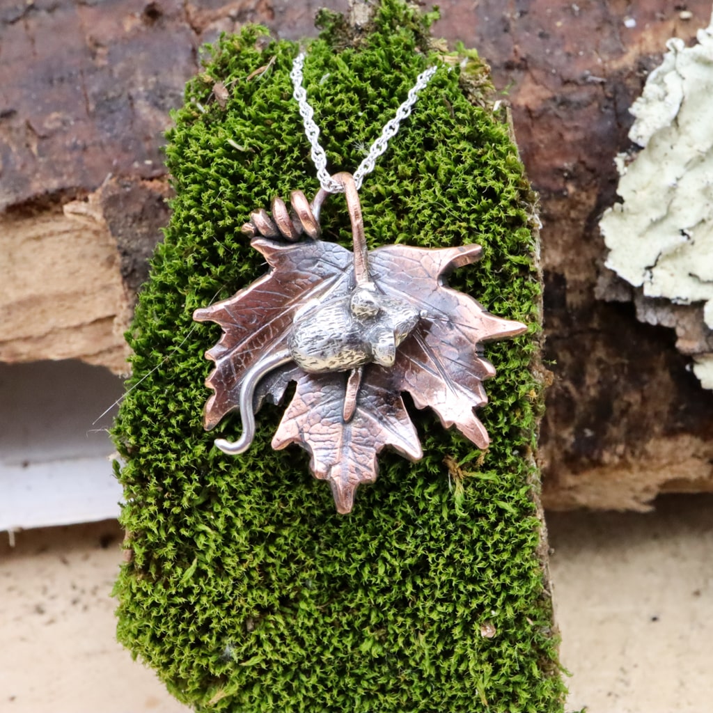 A hand carved sterling silver mouse sits in the middle of a handmade copper maple leaf. The necklace is about 1 inch tall and shown on a piece of dark green moss.