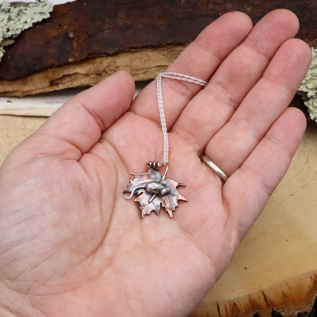 A hand is shown holding the tiny silver mouse on a maple leaf necklace for size reference.