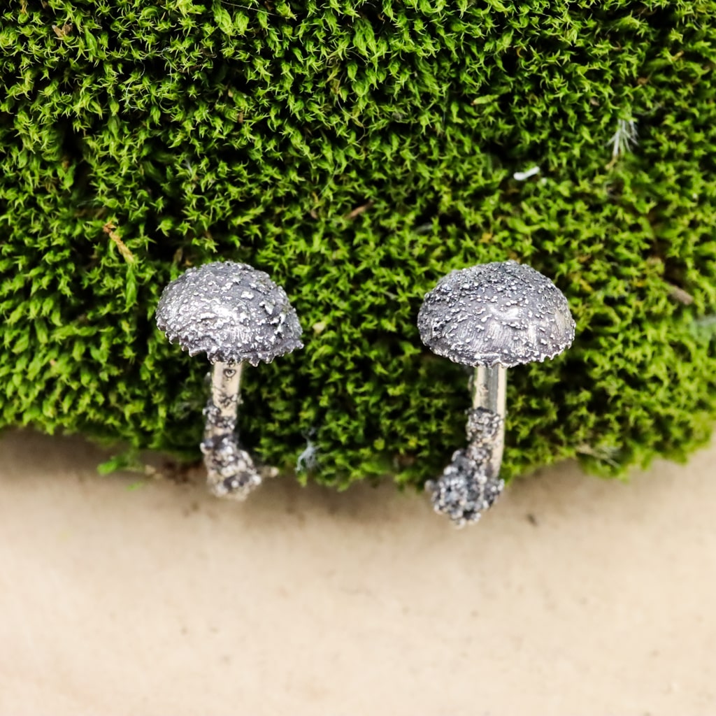 The tops of the silver mushroom earrings. They look realistic with the pattern on the tops. The earrings are shown on a piece of dark green moss.