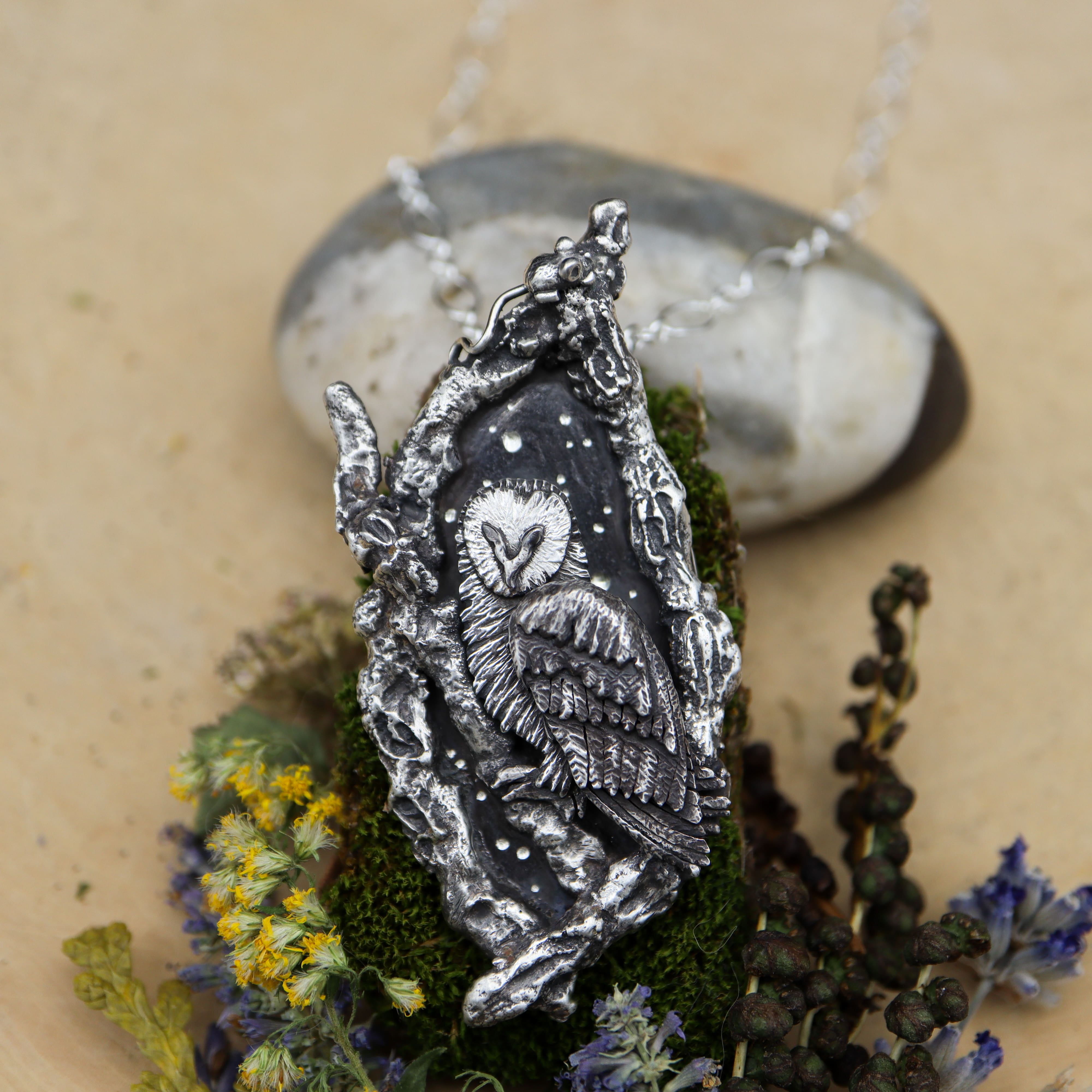 A handmade sterling silver own necklace is shown being surrounded by silver tree branches. At the top of the branches are a tiny hand carved silver mouse hiding. There are yellow and purple flowers at the base of the necklace.