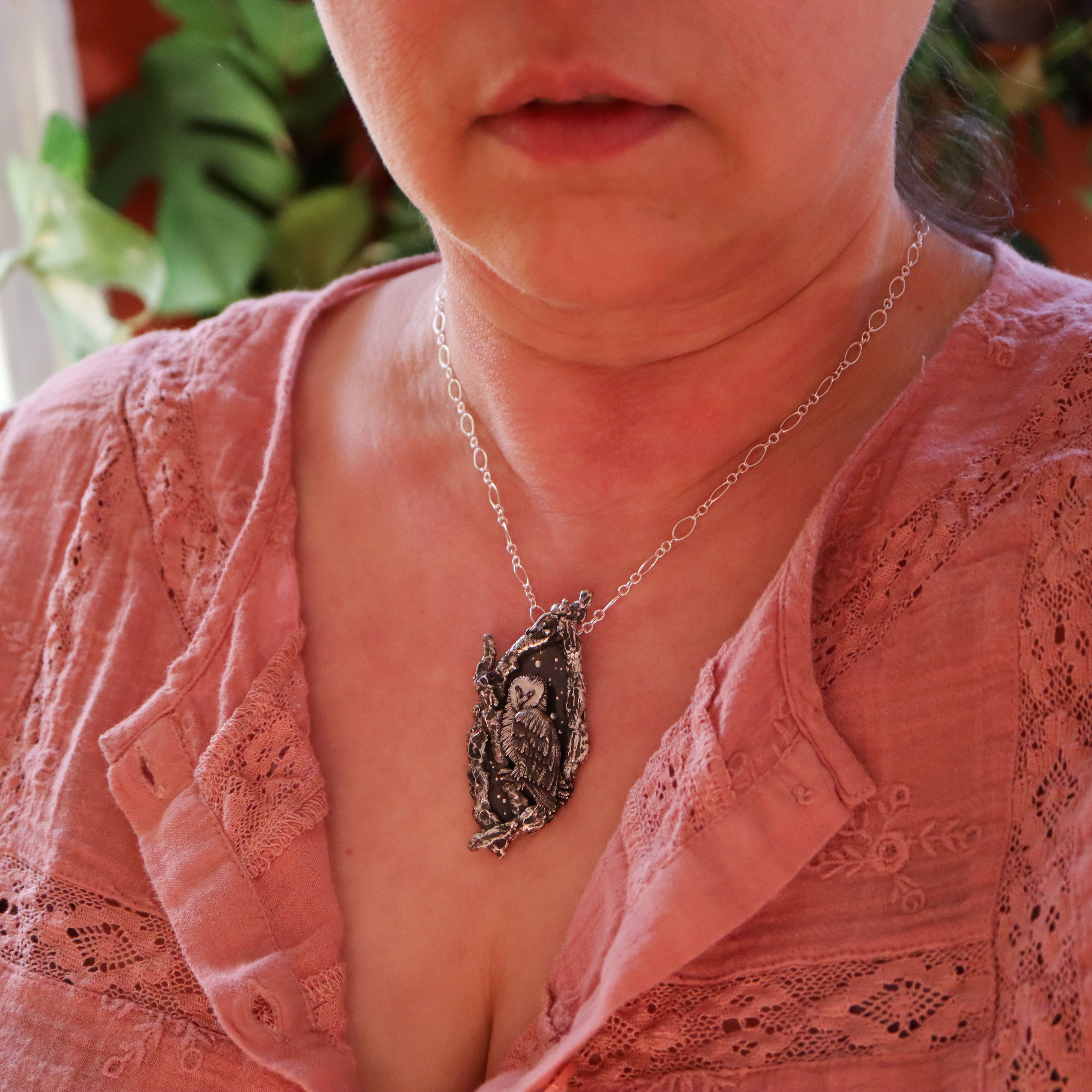 A woman is shown wearing the owl necklace. She is wearing a v neck pink lacy shirt.