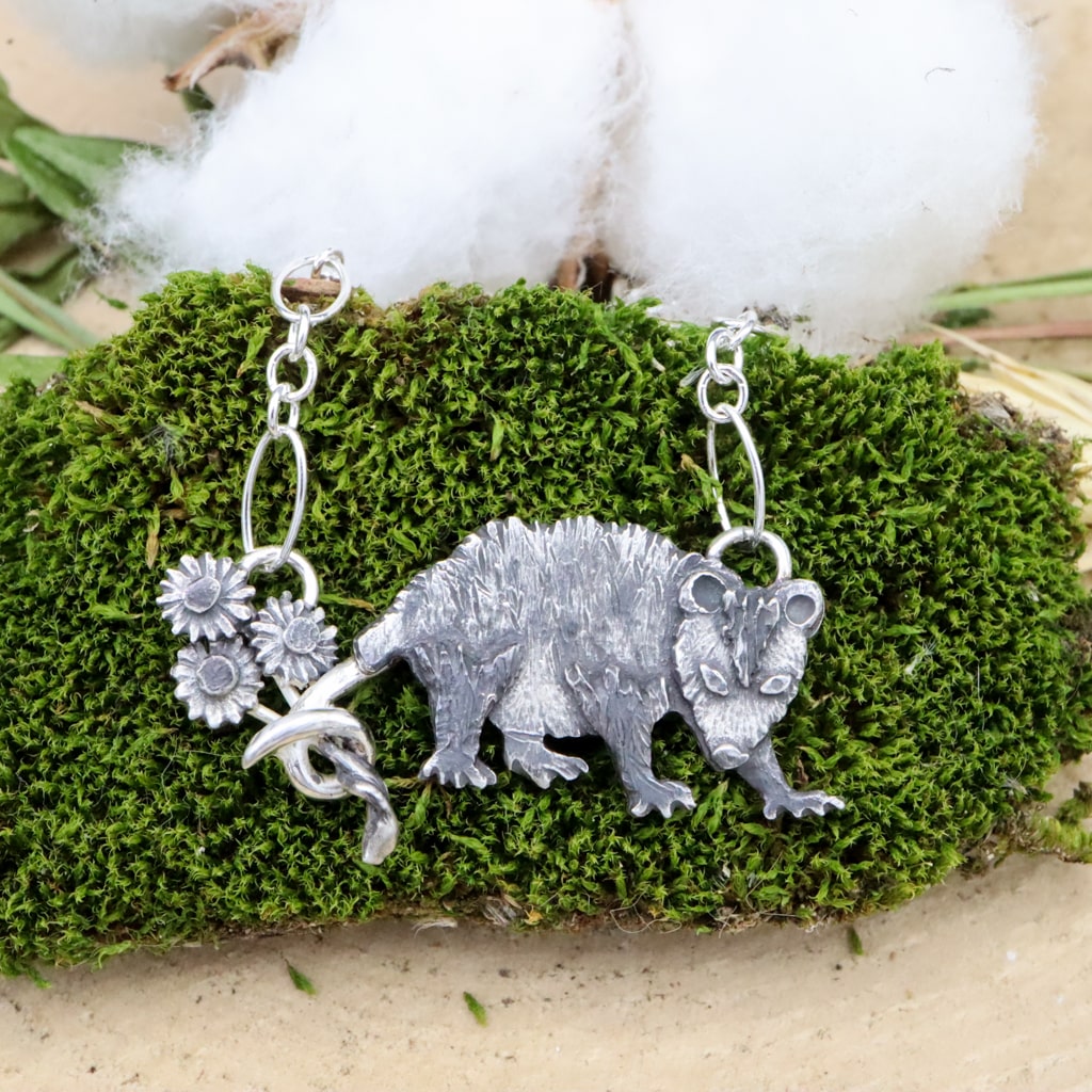 A sterling silver Virginia opossum necklace. She is shown holding a bouquet of three daisy flower stems in her curled up tail. The necklace is shown on top of a dark green piece of moss.