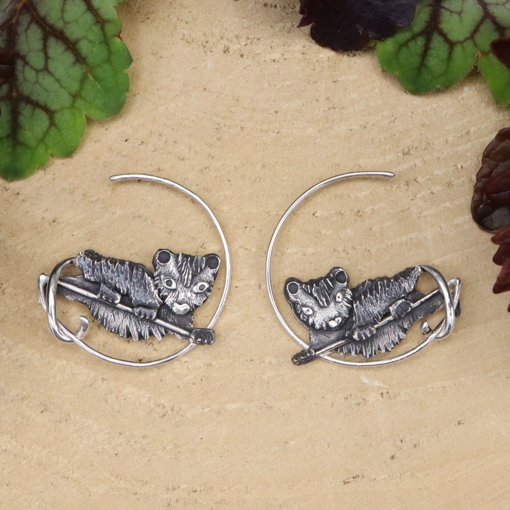 Sterling silver handmade hoop earrings featuring baby opossums in the center of them. The earrings are about 1 inch tall and 1 inch wide. they are shown with some green leaves above them and on a light tan piece of wood.