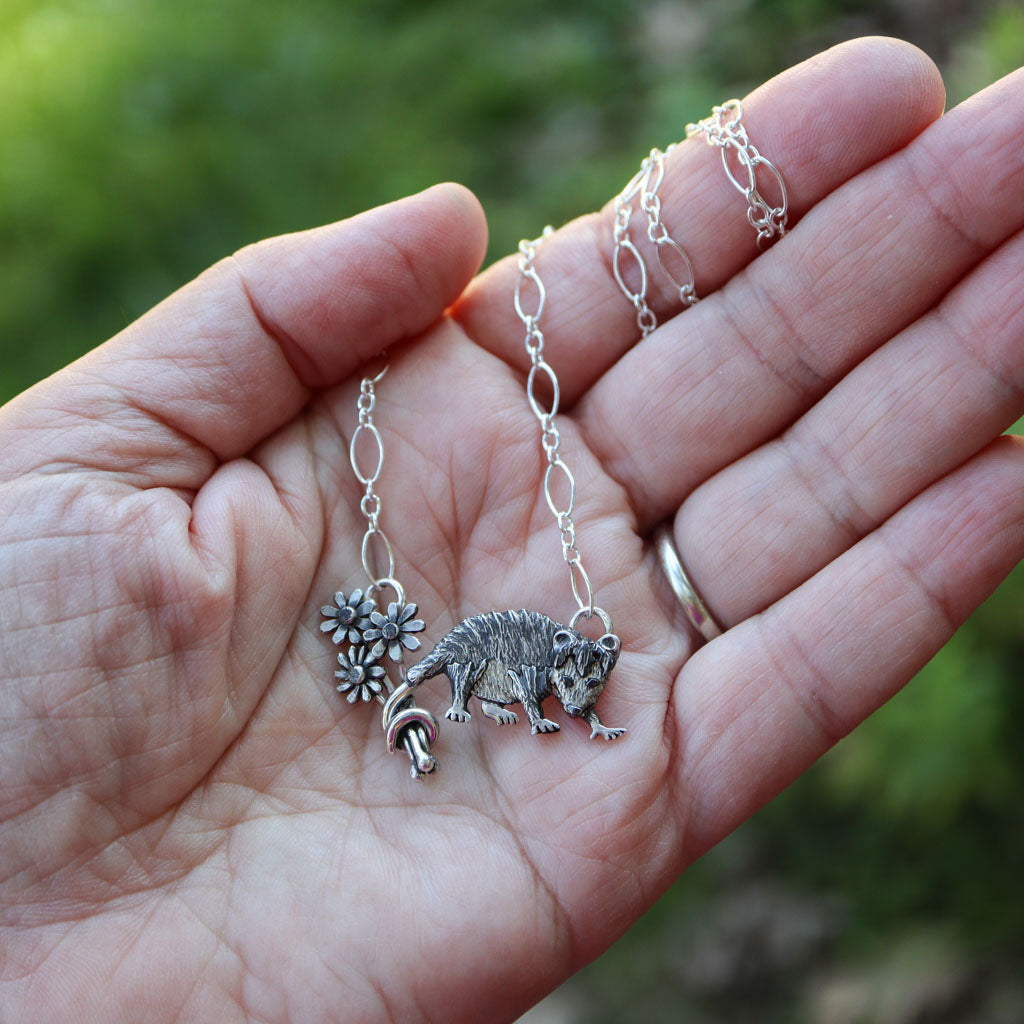 A close-up of a hand holding a Virginia opossum holding a bouquet of daisies in her tail, with a blurred green outdoor background.