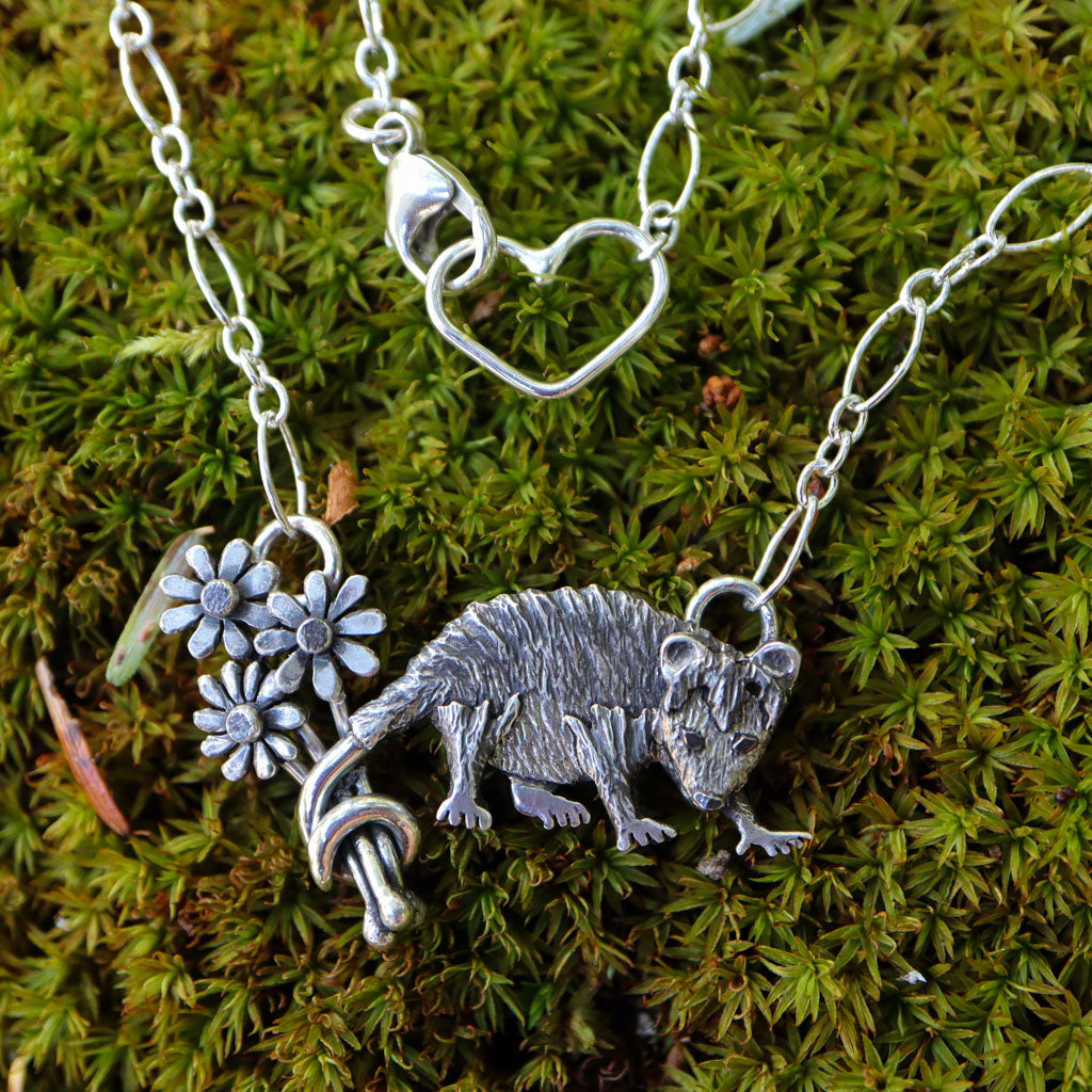 A sterling silver pendant necklace featuring a Virginia opossum holding flowers in her wrapped tail. She is shown on a bed of green moss.