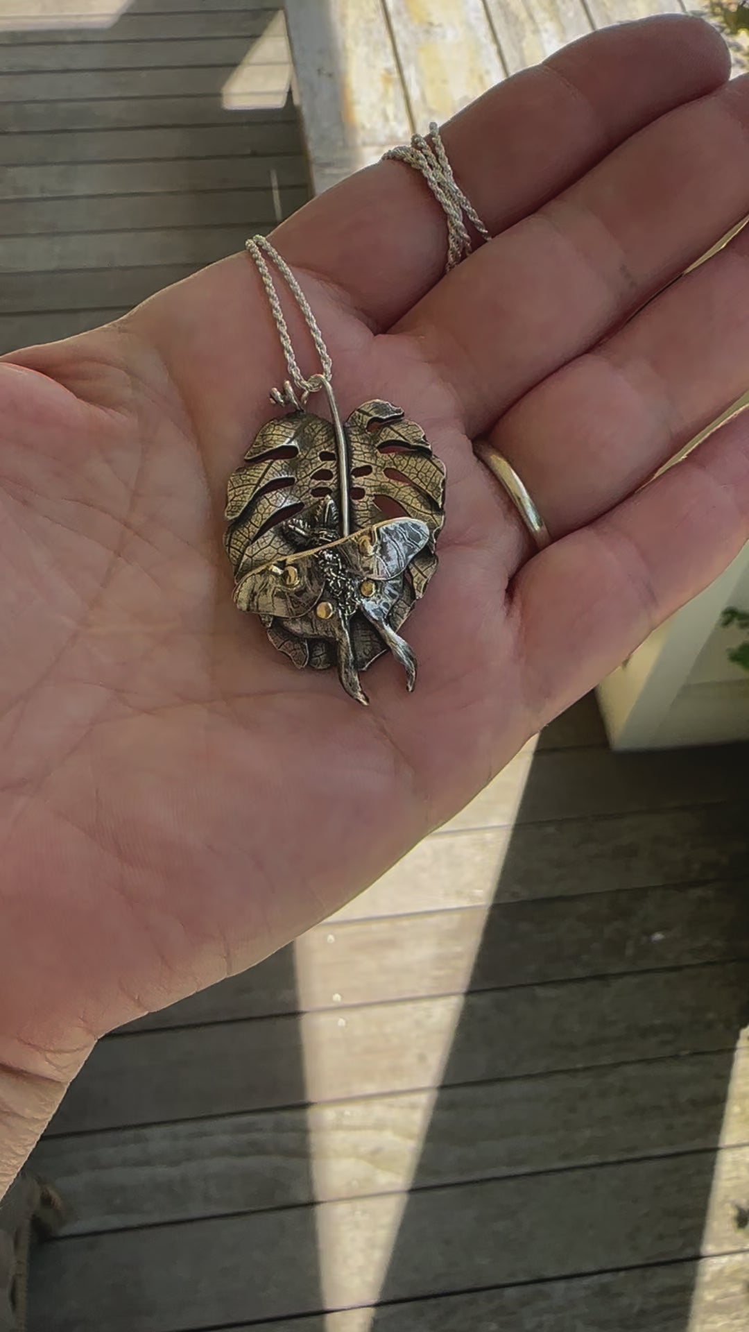 A handmade sterling silver and gold luna moth sits upon a monstera deliciosa leaf necklace. A hand is holding the necklace and showing it at different angles. It is about 1.75 inches tall.