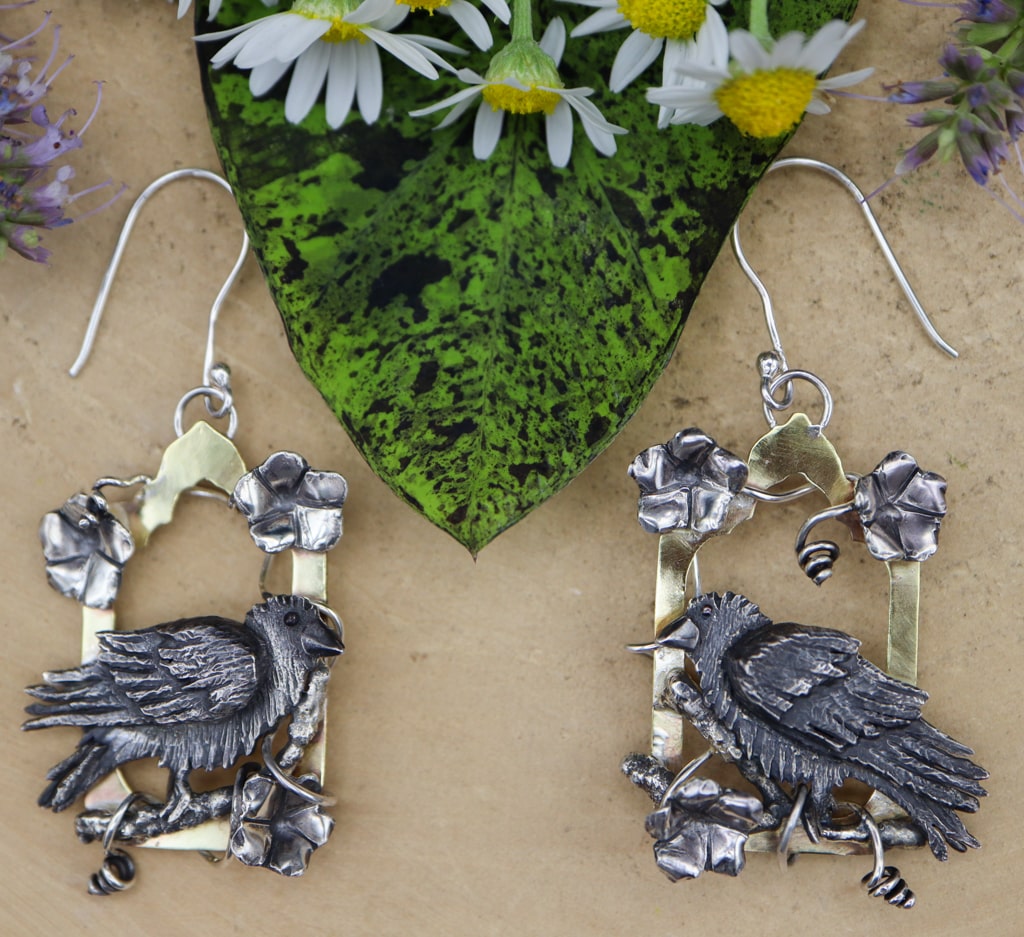 Sterling silver and brass earrings featuring fluffy black ravens surrounded by vines of moonflowers. Each earring is a little different from the other one. They are about 2.5 inches long and shown on a light tan piece of wood with flowers around.