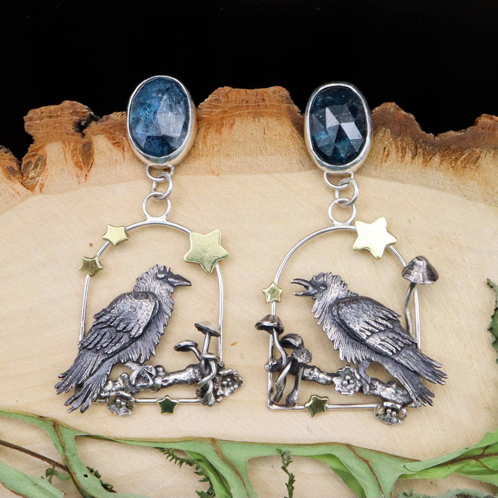 Handmade sterling silver raven mismatched earrings. Each earring features a hand carved silver raven bird sitting on a tree branch with crab apple blossoms around them. There are bright brass stars and mushrooms growing around them. At the top of the earrings there are dark blue moss kyanite gemstones.