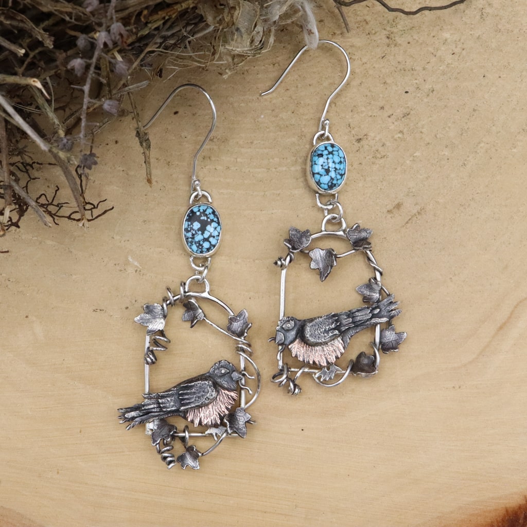 A pair of American robin earrings. They are sitting in a window shape and surrounded by vines. There are two light blue turquoise stones at the top of the earrings. They are shown on a light tan piece of wood.