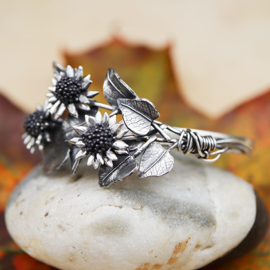 A sterling silver sunflower bouquet bracelet is shown on a white stone with fall leaves in the background.