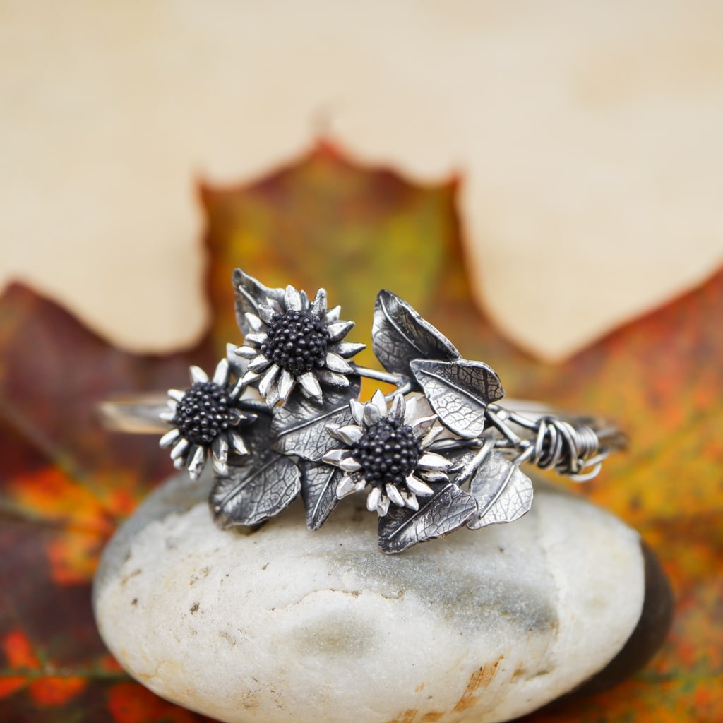 The front view of the three sunflower stems that are on the sterling silver bracelet. There are fall leaves in the background and it is shown on a white stone.