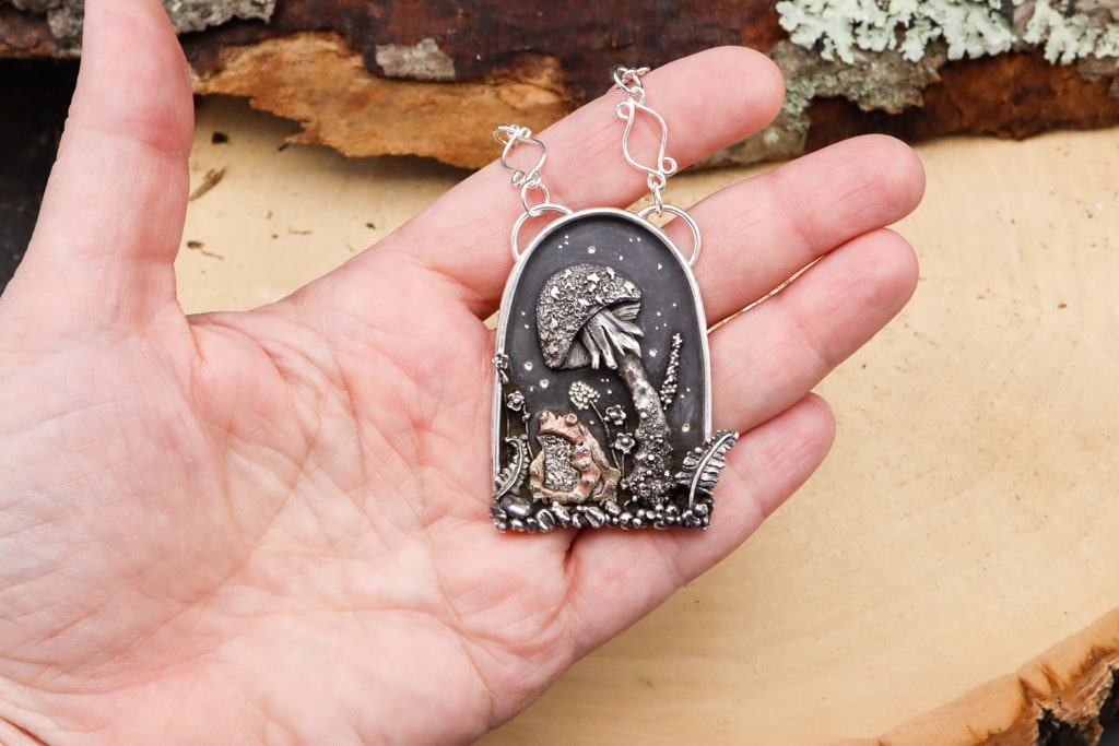 A hand is shown holding the toad pendant to show its size. The pendant is quite large at about 3 inches tall.